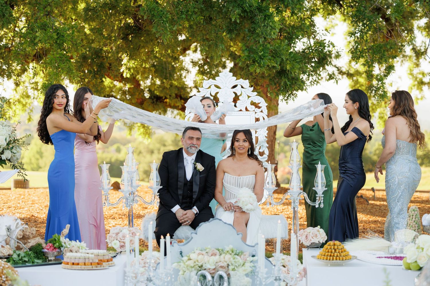 Elegant outdoor wedding reception with tables decorated in white linens and floral centerpieces under sunlit trees.