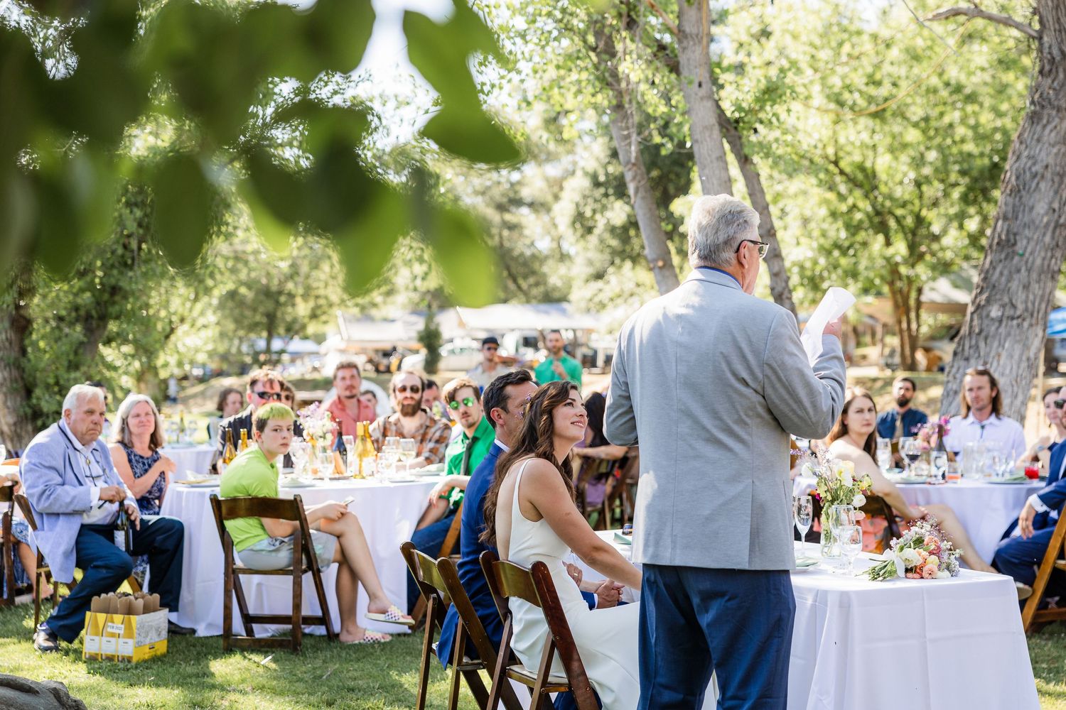 Wedding guests mingle and socialize at an outdoor reception under trees on a sunny afternoon.