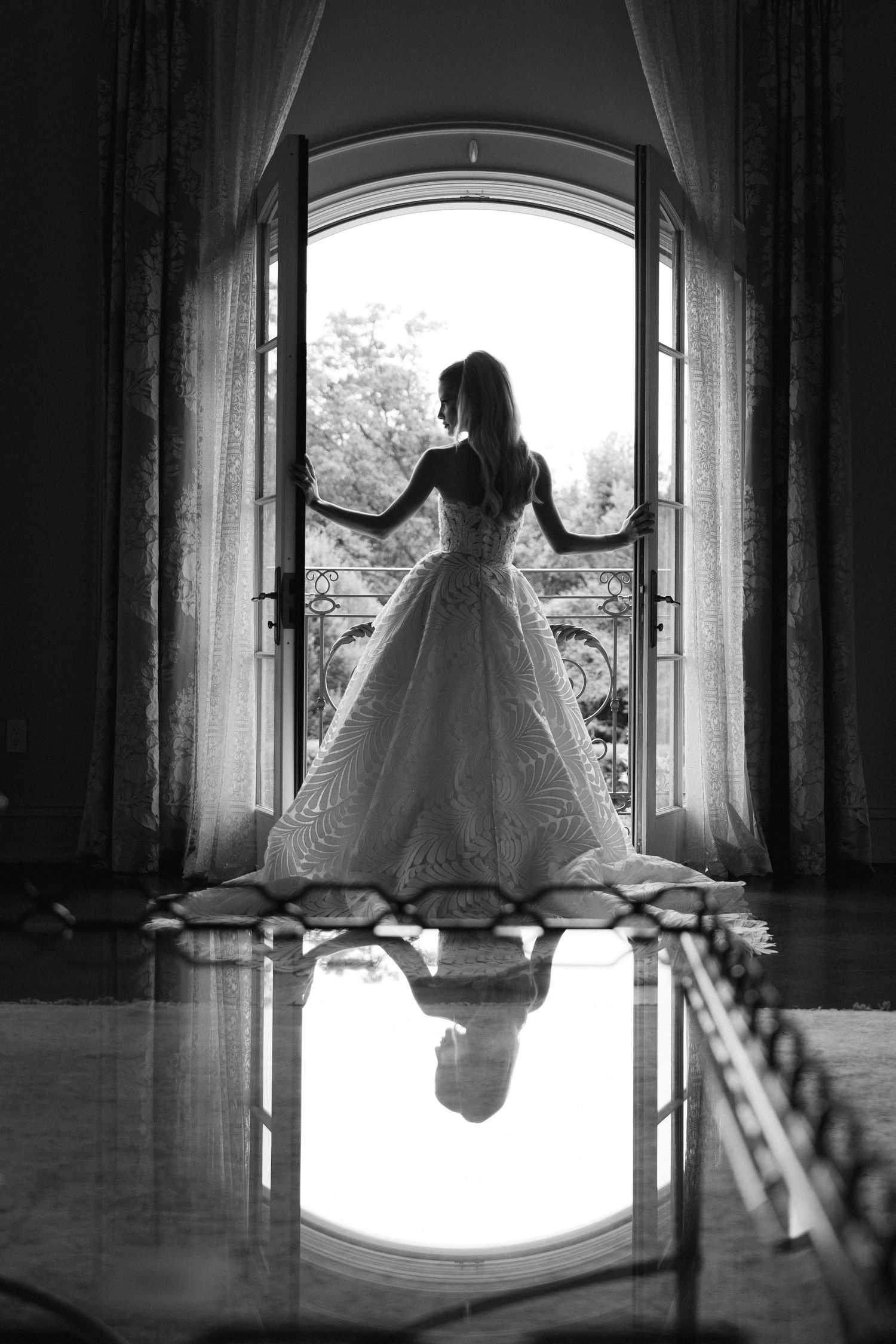 A silhouetted figure in a wedding gown stands in an arched doorway with their reflection mirrored on the polished floor.