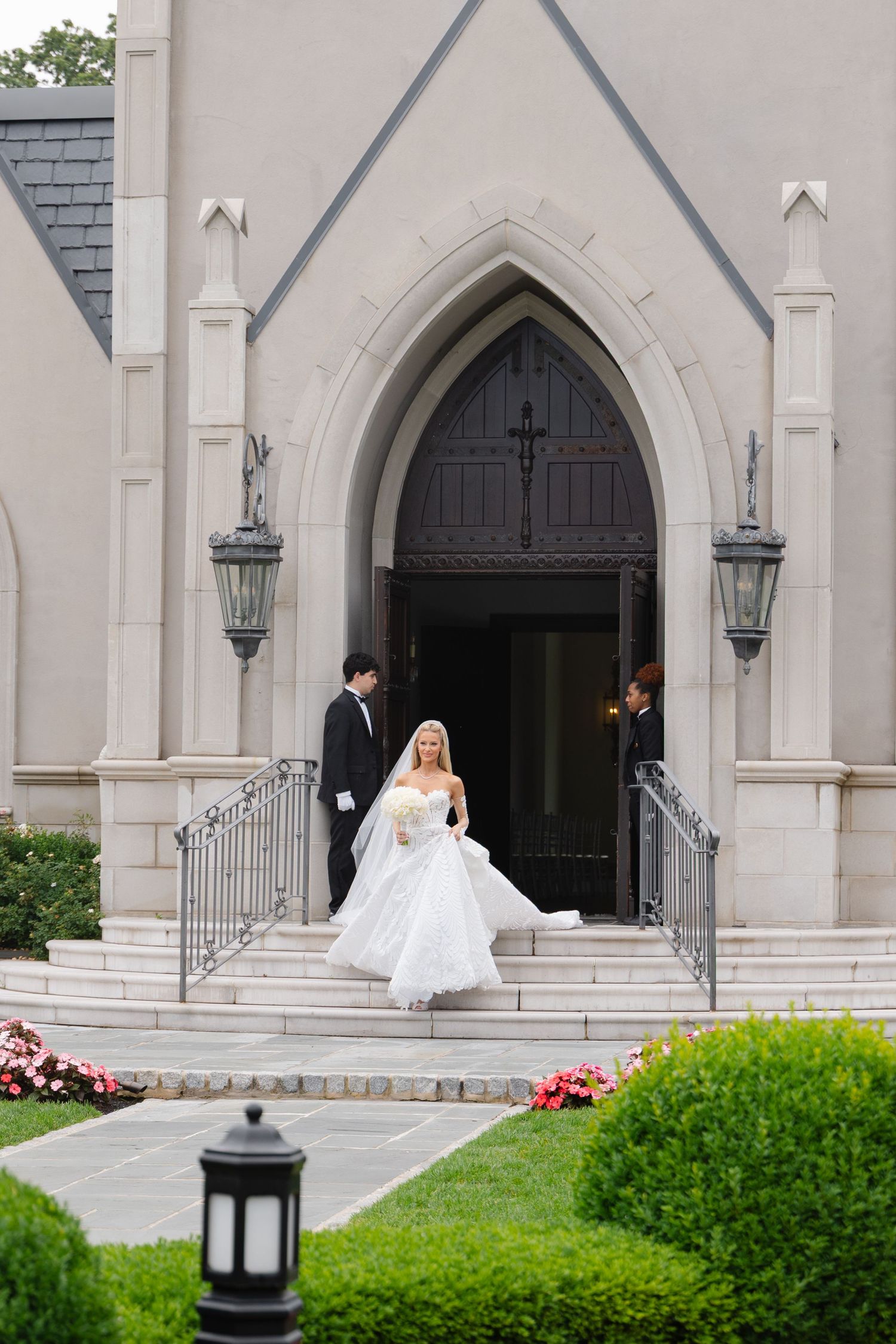 A couple in formal wedding attire stands outside a white stone church with gothic arched doorways and lanterns.