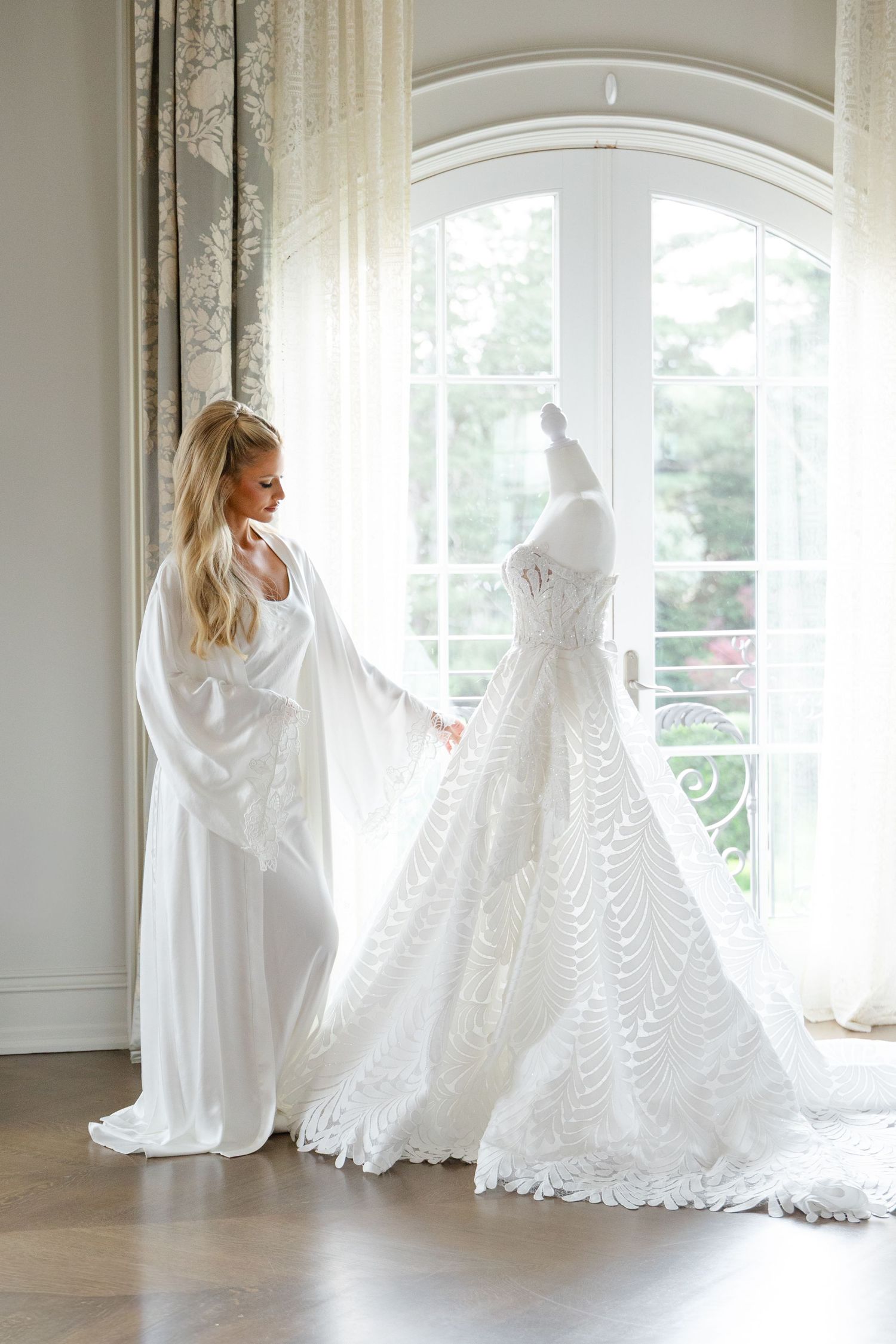 A figure in a white robe admires a flowing lace wedding dress displayed near bright windows in an elegant room.