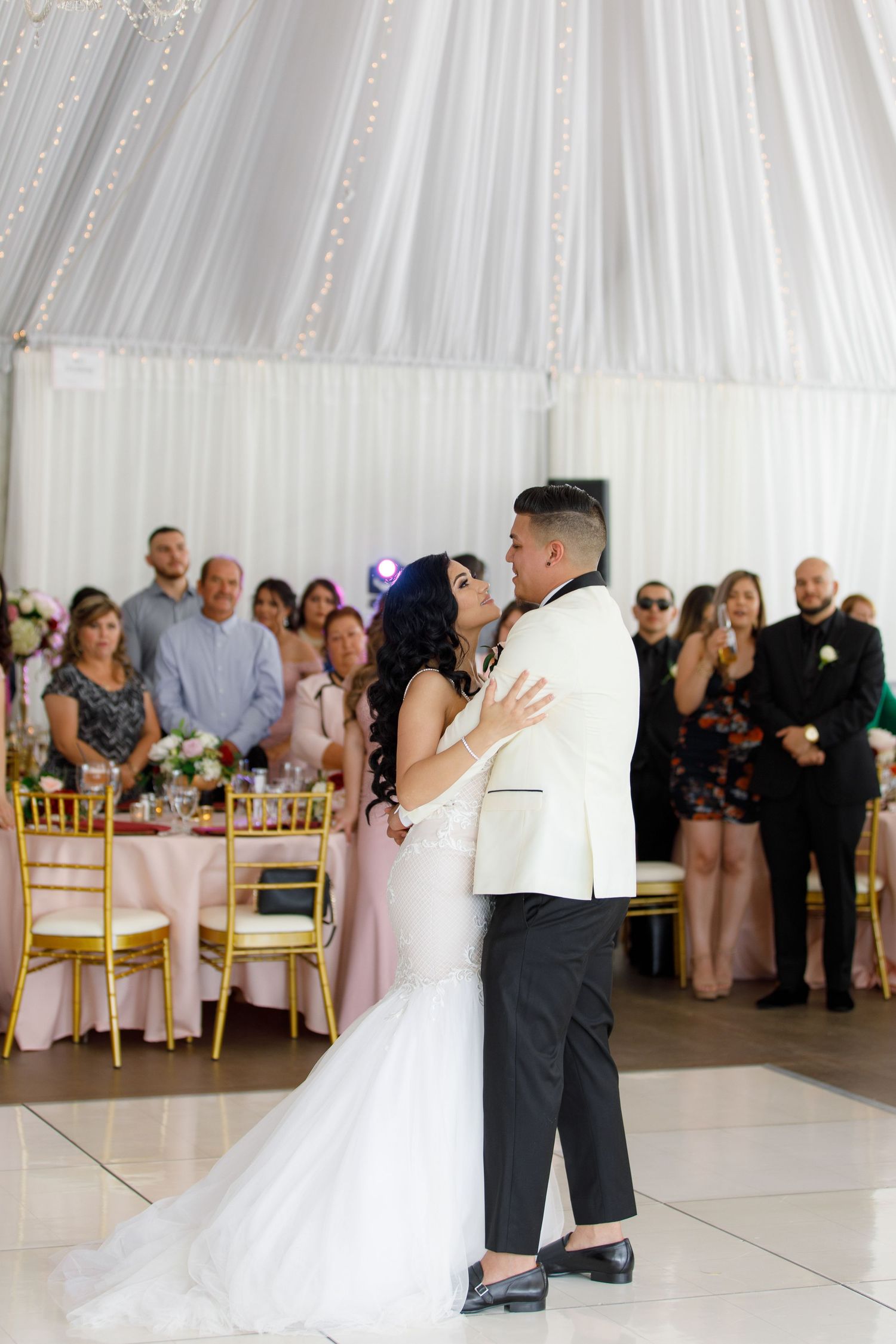 A couple shares their first dance at a wedding reception in a white-draped tent venue with guests watching from their tables.