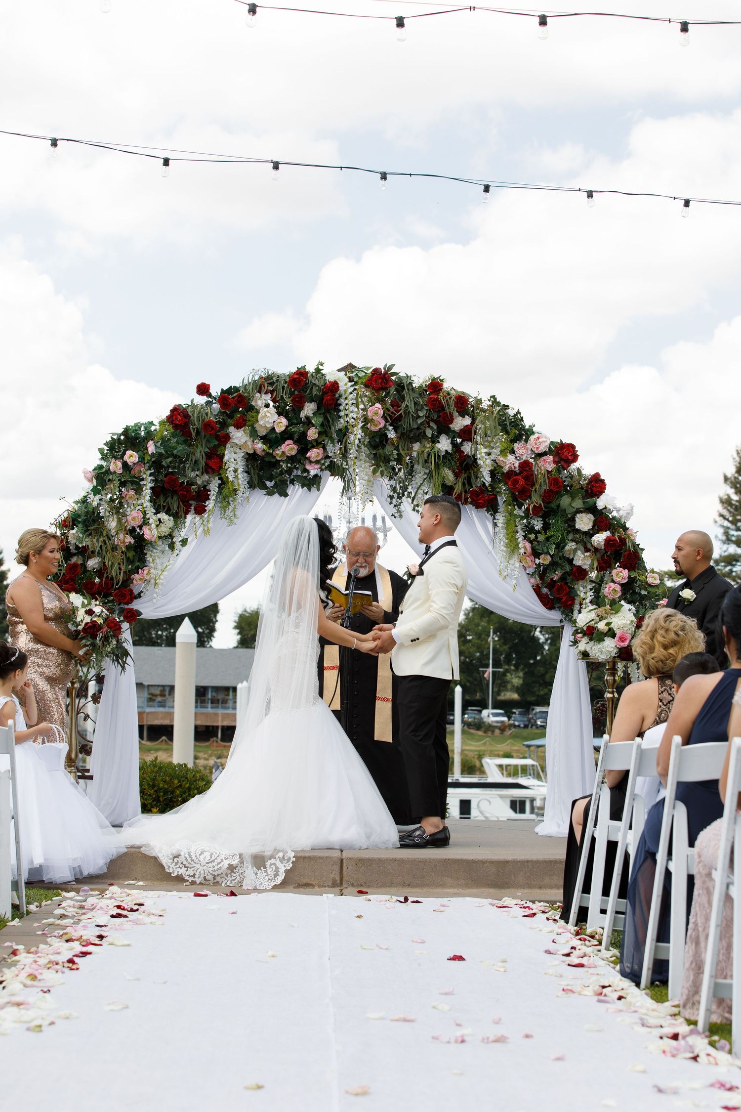 Outdoor wedding ceremony setup with white fabric arch adorned with red and white flowers and scattered rose petals.