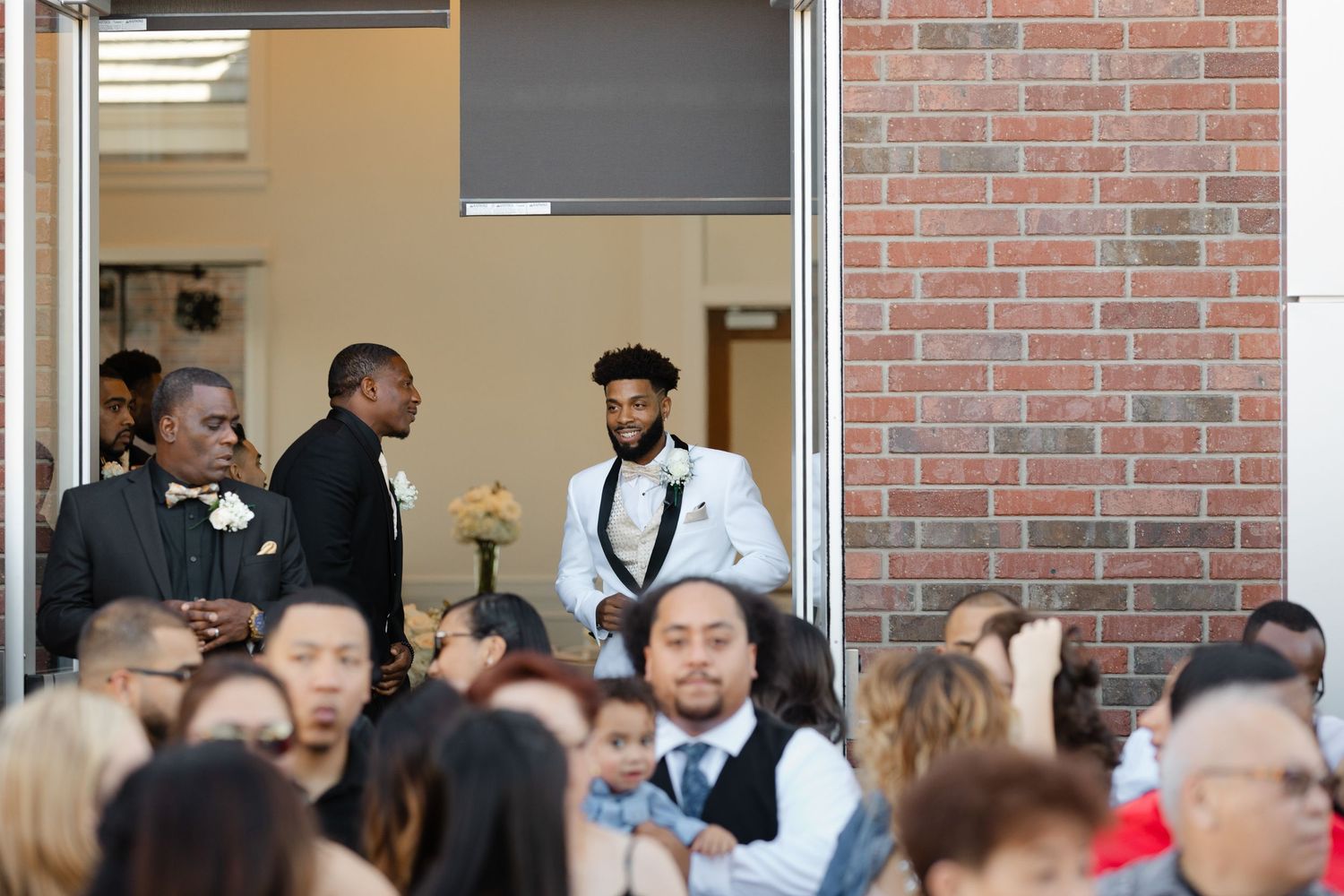 A crowd of people gathered at an indoor event with brick walls and formal attire visible in the background.