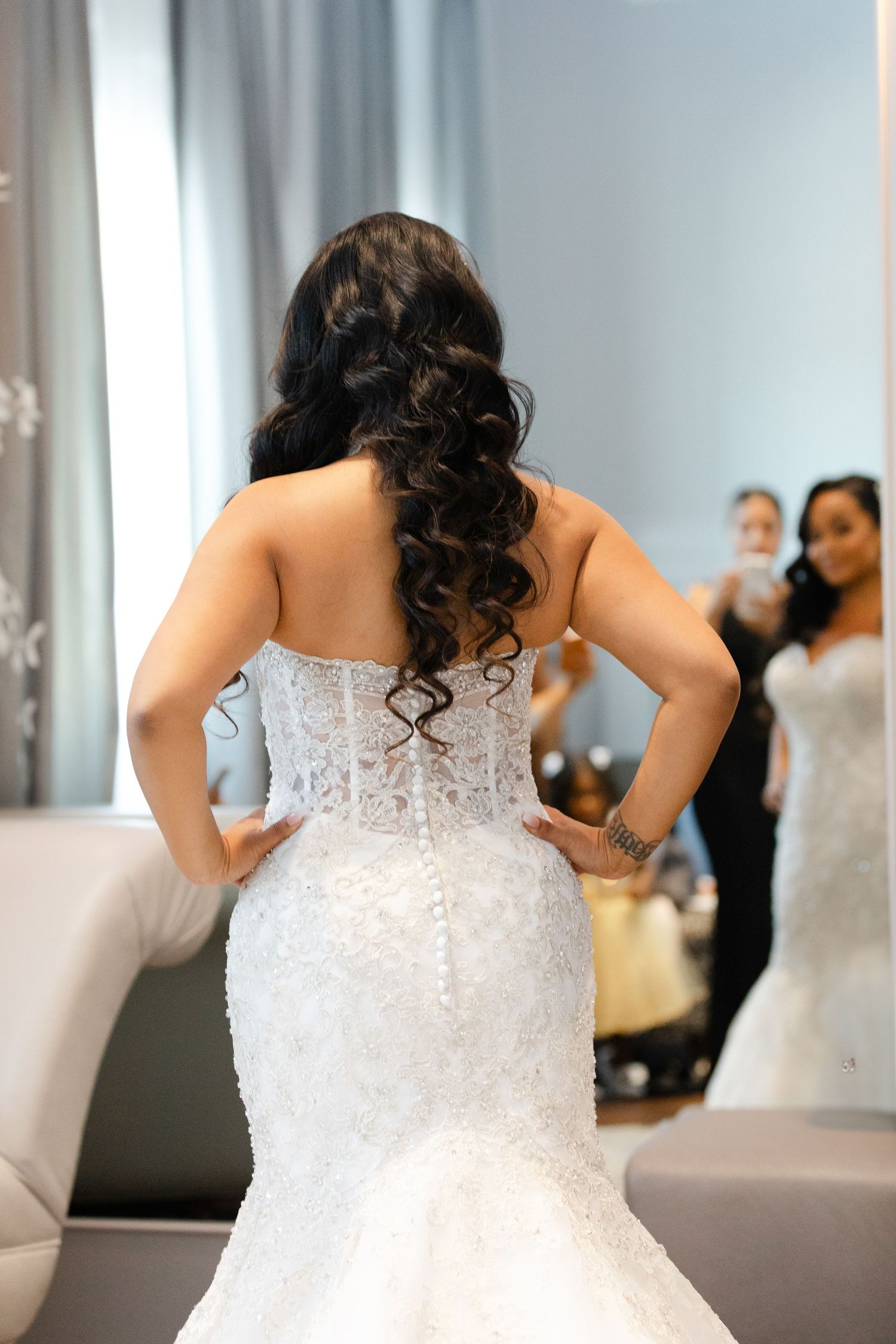Back view of a bride in a strapless white wedding dress with lace details being fitted in a bridal boutique.