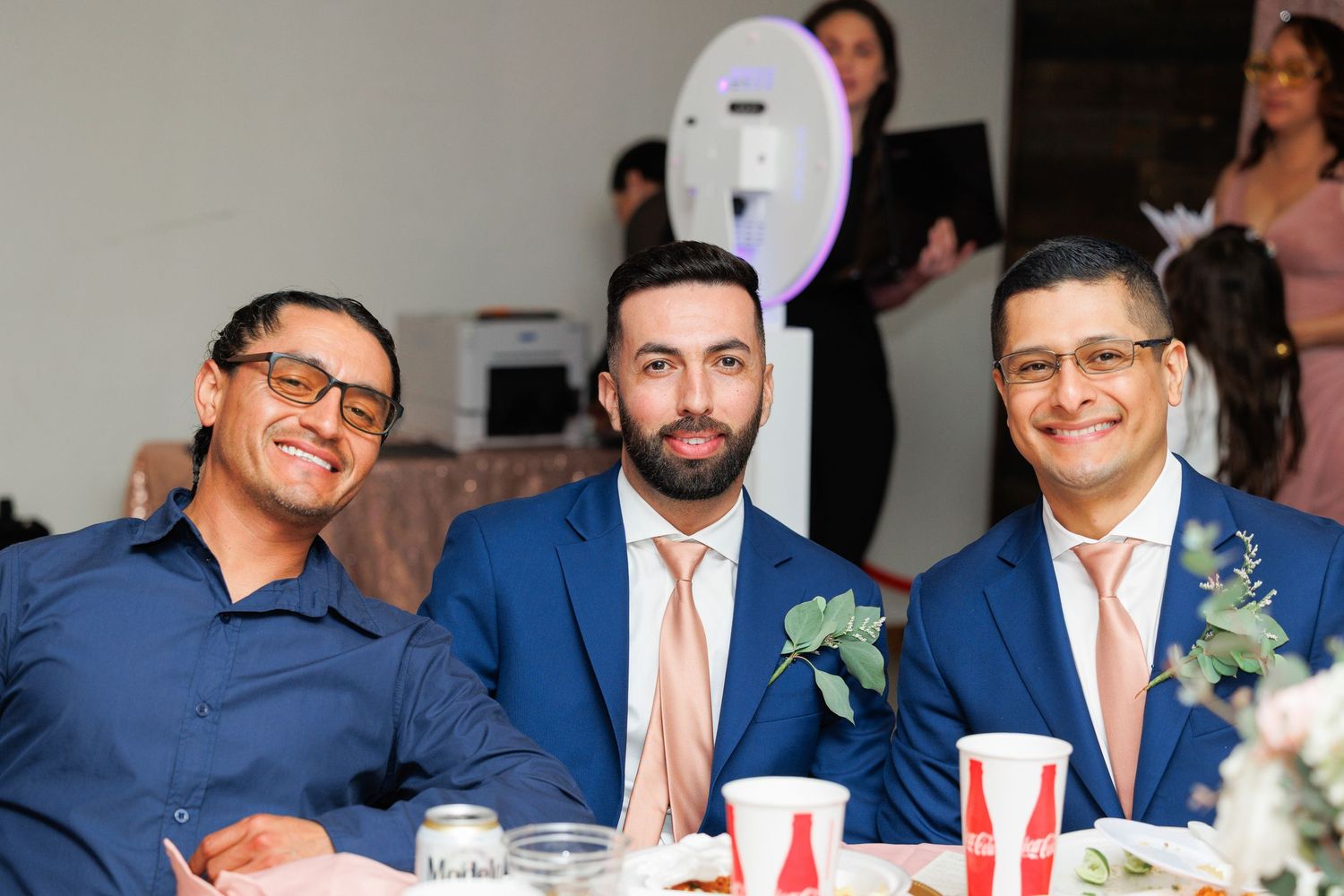 A group of wedding guests in navy suits and pink ties share laughs and drinks during a reception celebration.