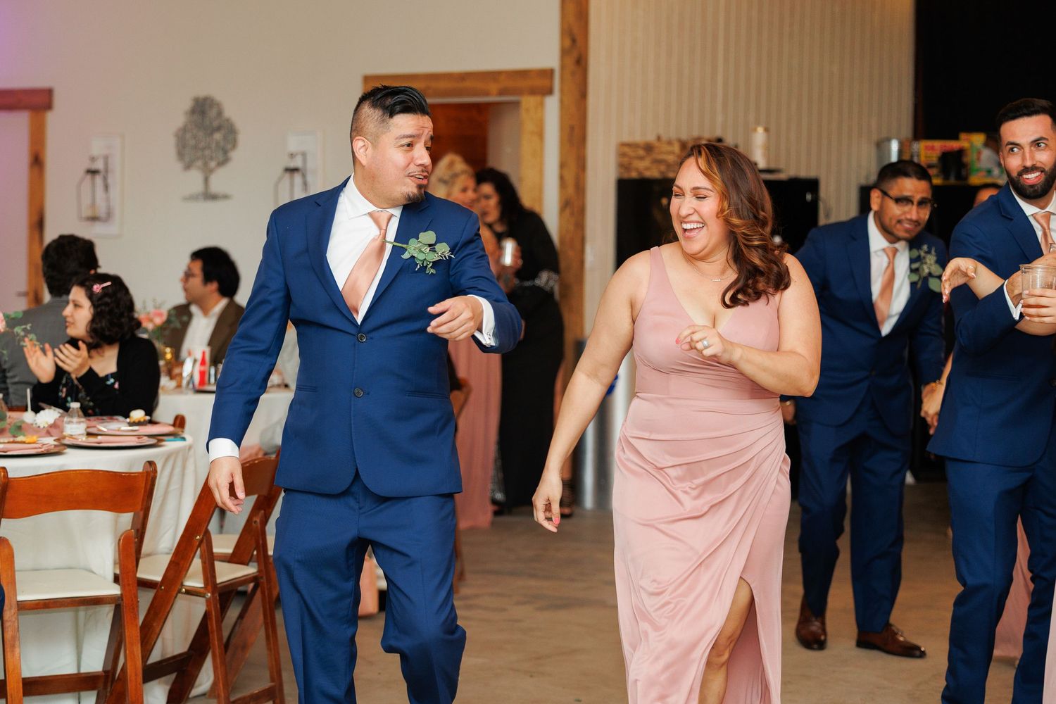 Wedding party in navy suits and blush pink dresses dance and celebrate during a reception.