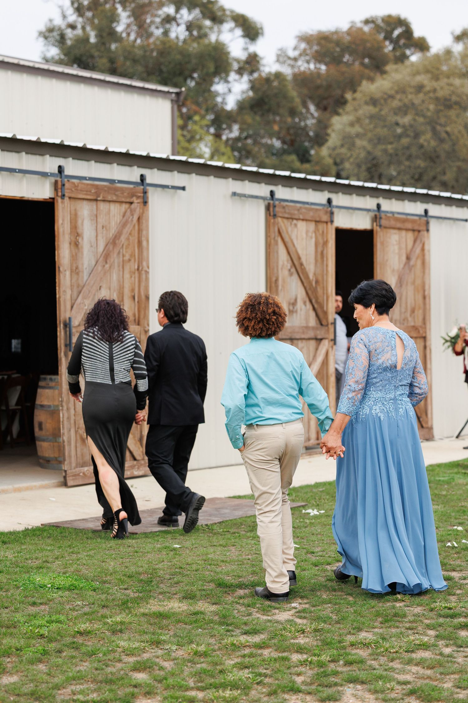 A group walks together in a line toward rustic wooden barn doors on a grassy path at an outdoor venue.