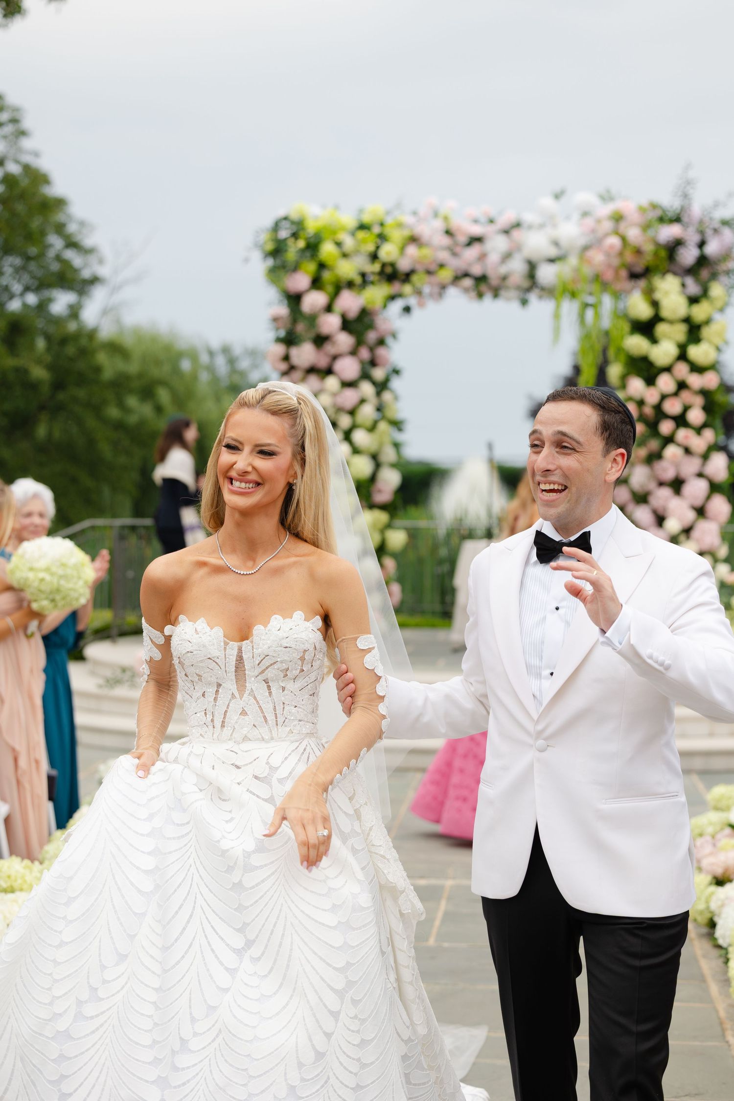 A joyful wedding celebration with the couple sharing laughter and smiles surrounded by pink and white floral arrangements.