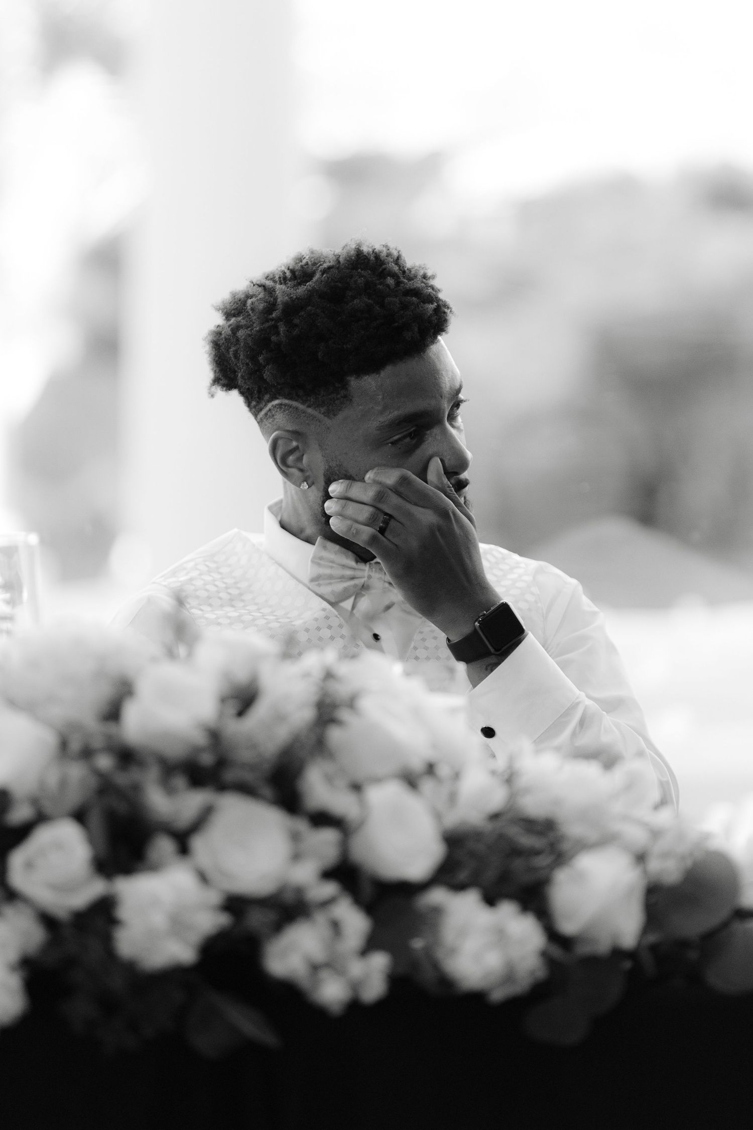 Black and white photo of someone with curly hair sitting behind white roses with their head in their hands.