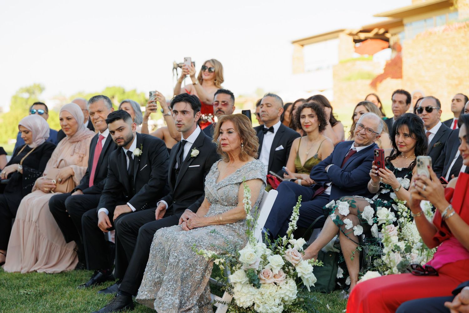 Wedding guests seated outdoors at a ceremony with floral arrangements and golden hour lighting.