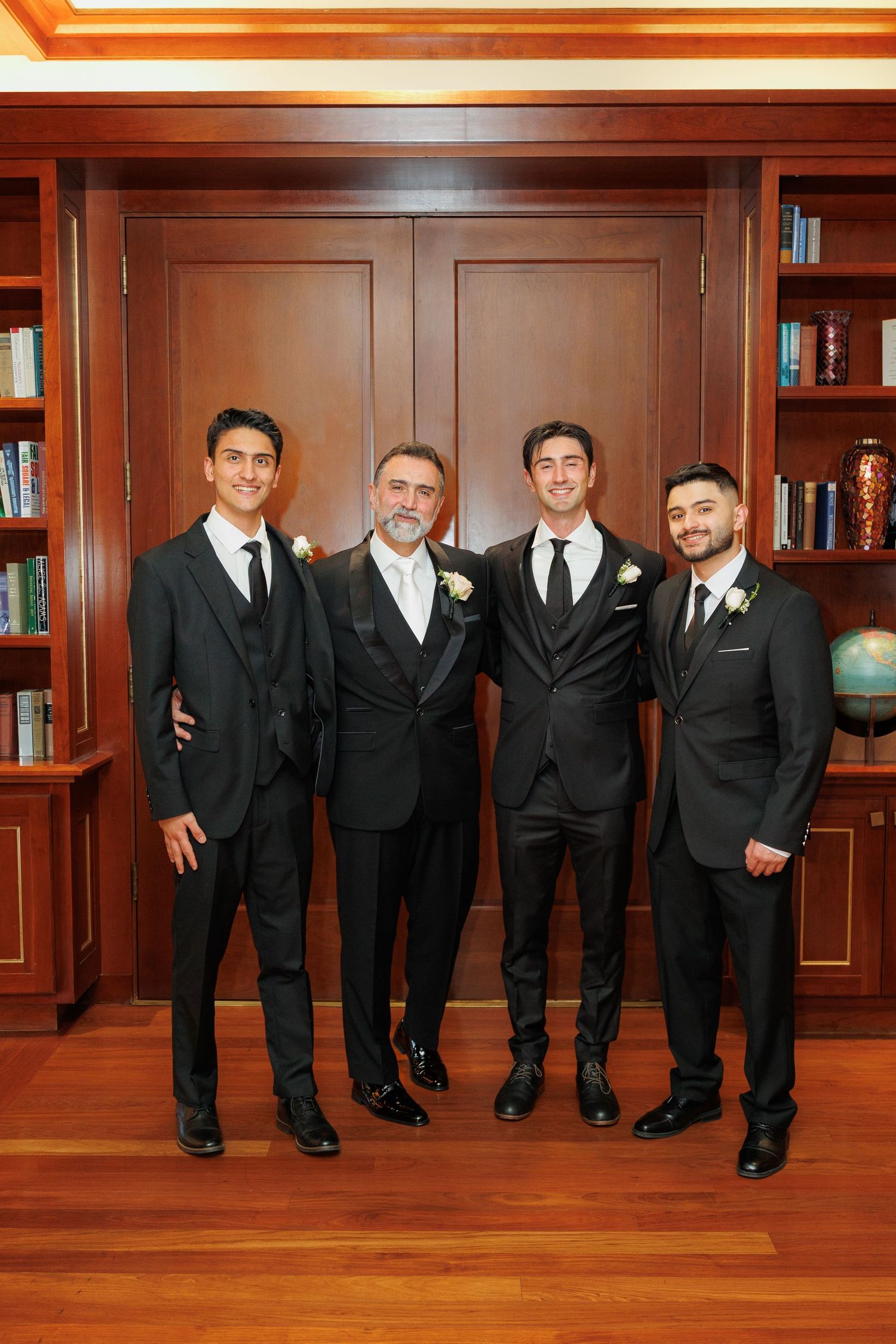 Groomsmen in black suits pose playfully in a wood-paneled library room for a fun wedding photo.