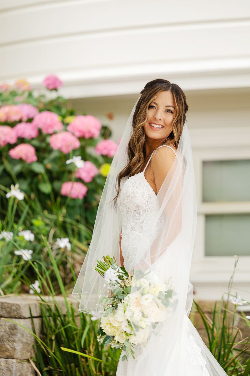 A bride in a white lace dress poses with her bouquet against pink hydrangeas and white architecture.