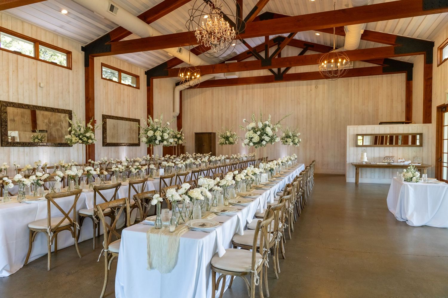 Rustic wedding reception venue with exposed wooden beams, chandeliers, and long tables set in white linens.
