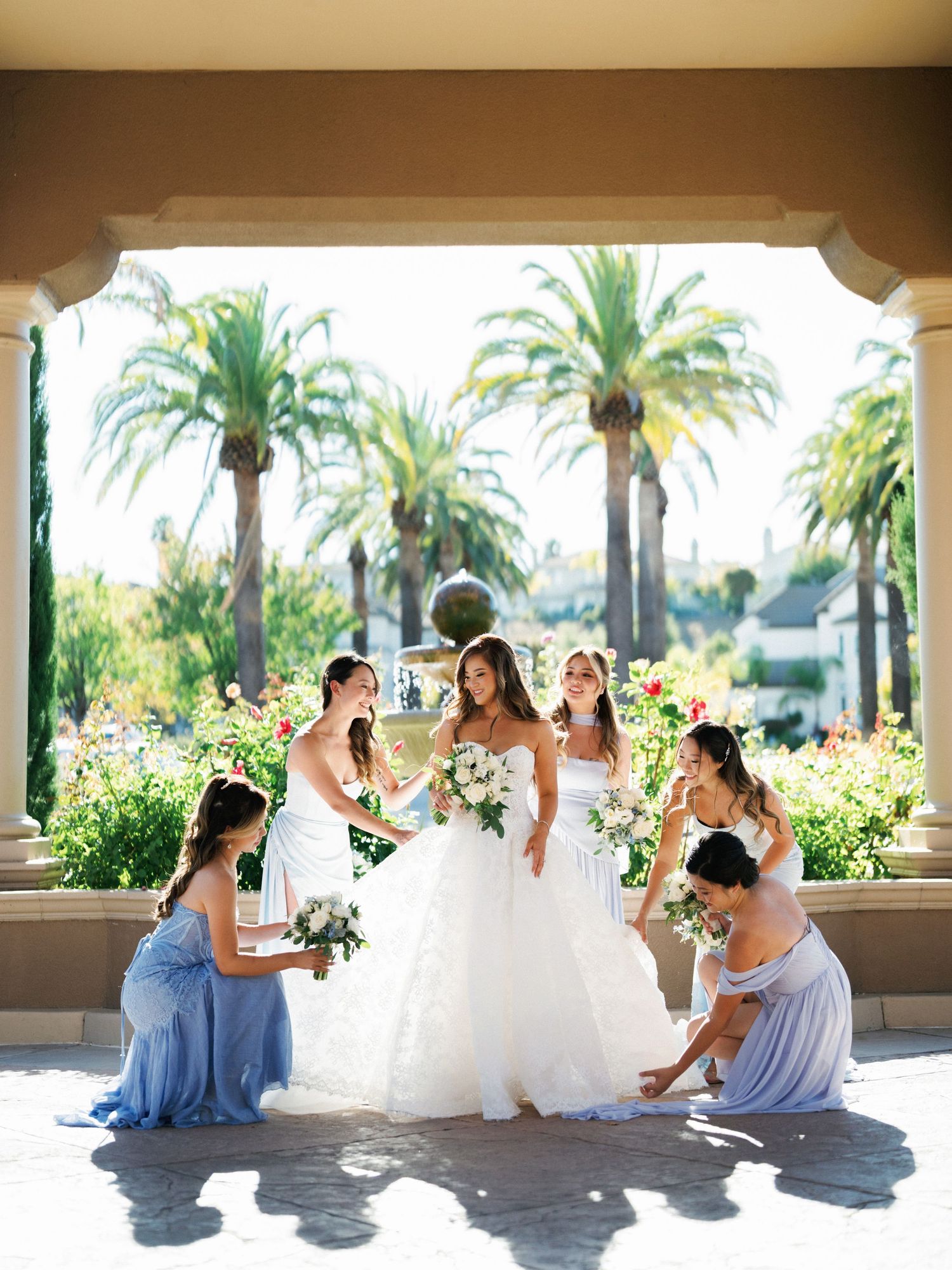 Wedding party in light blue dresses and white gown posing under a palm tree lined veranda.