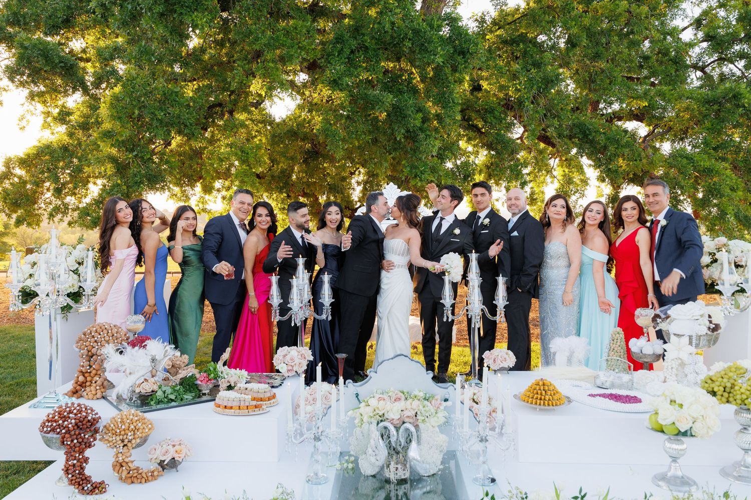 Wedding guests gather outdoors for a garden reception celebration under lush green trees at sunset.