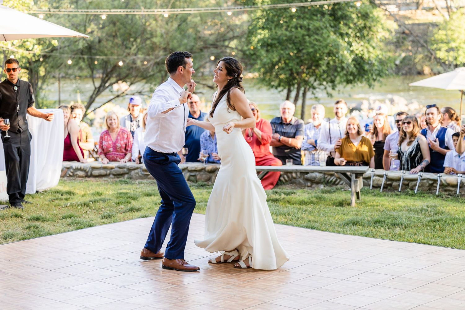 Guests dance together on a patio during an outdoor wedding reception with white chairs and trees in the background.