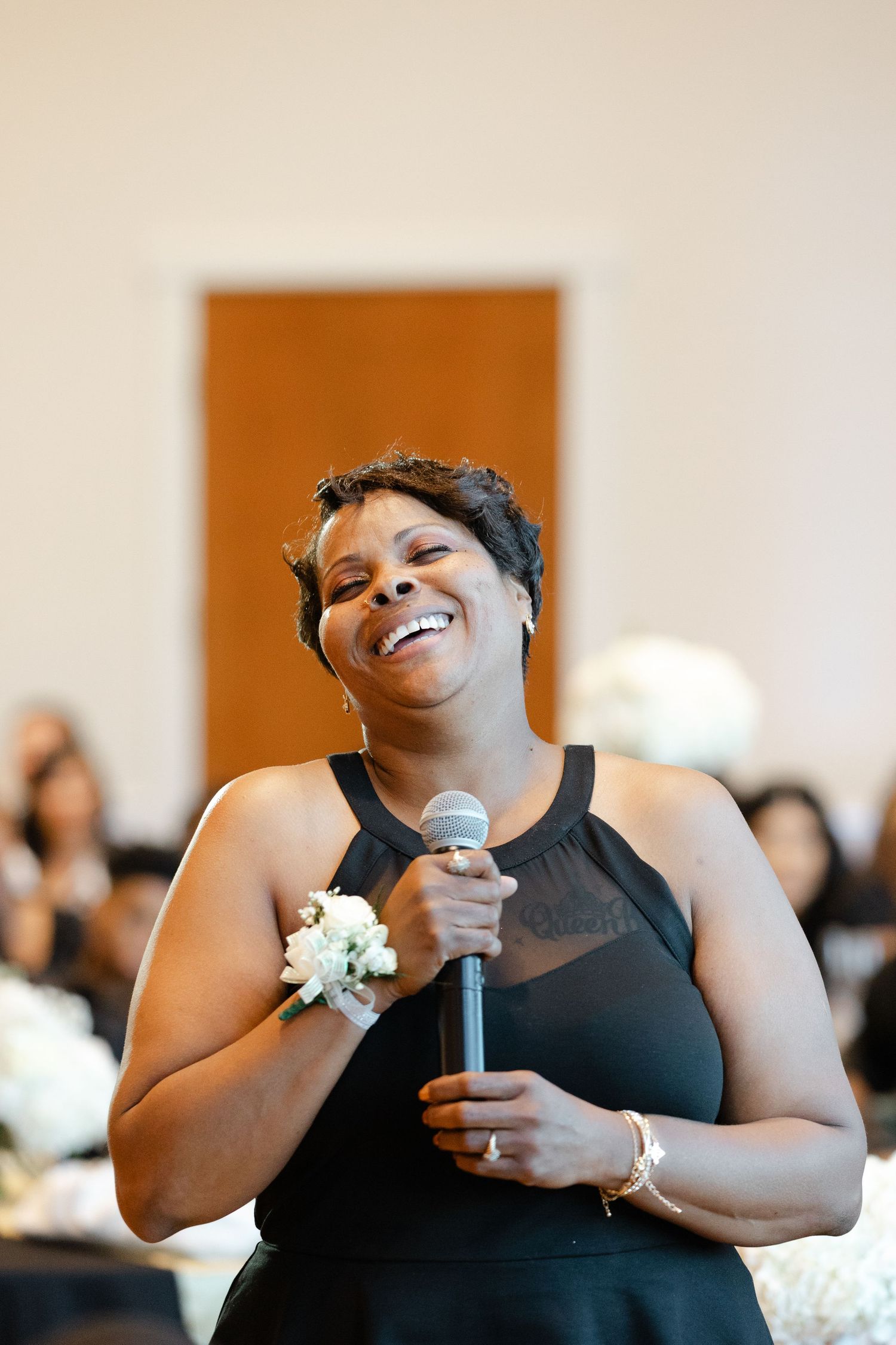 A person wearing a black dress gives a joyful speech at a formal event, with a white corsage on their wrist.