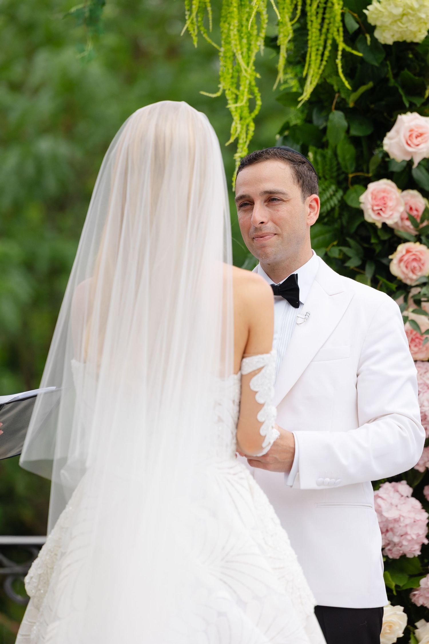 A romantic wedding ceremony moment with a bride in a flowing veil and groom in white under floral decor.