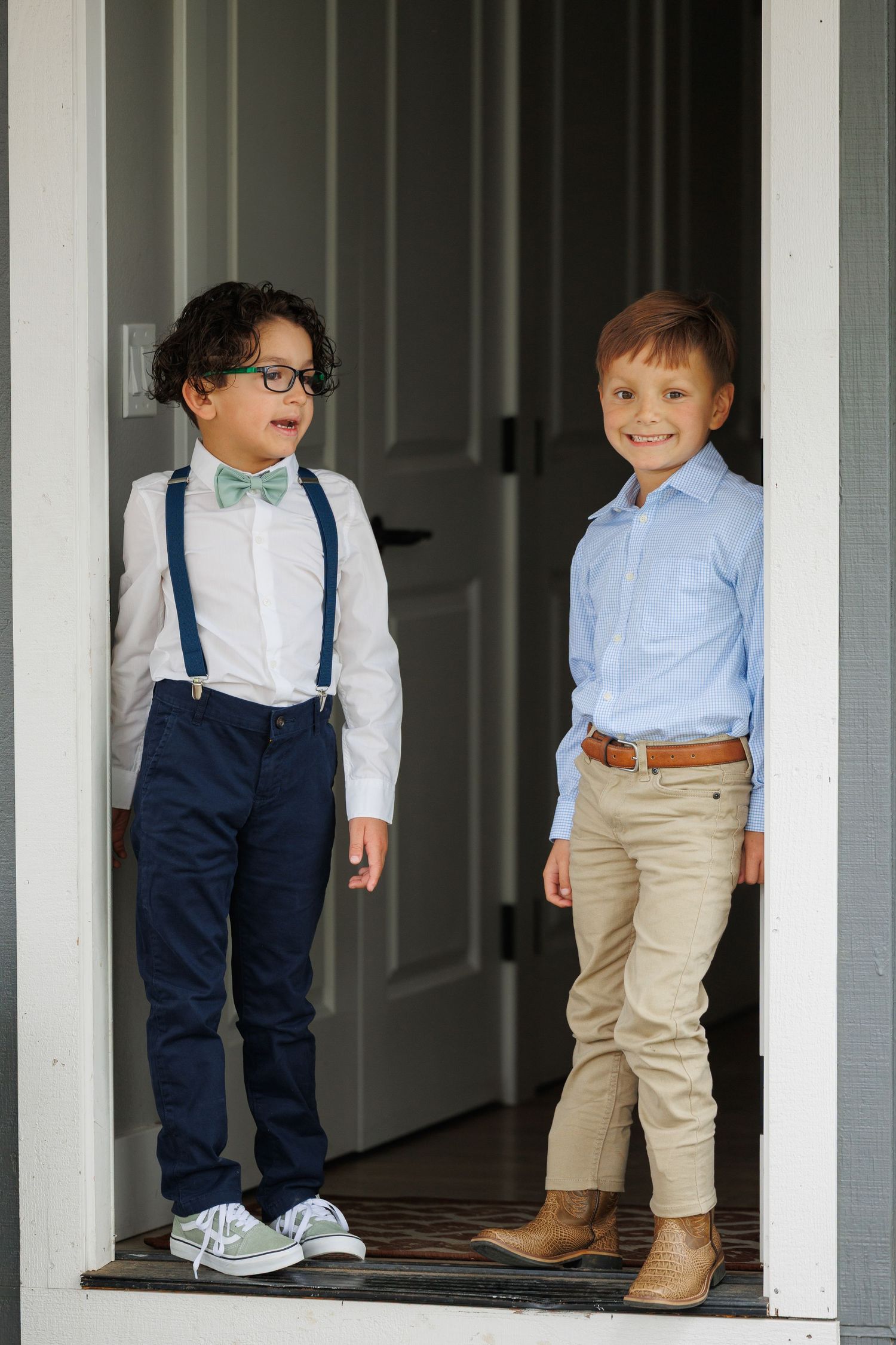 Young children in matching navy suspenders, khaki pants and white shirts stand on a porch with white columns.