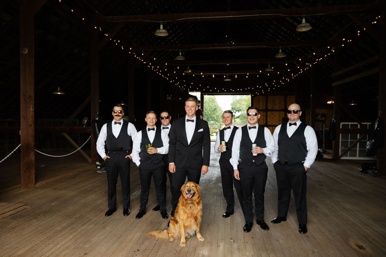 Wedding party in formal black and white attire poses with golden retriever in rustic indoor venue with string lights.