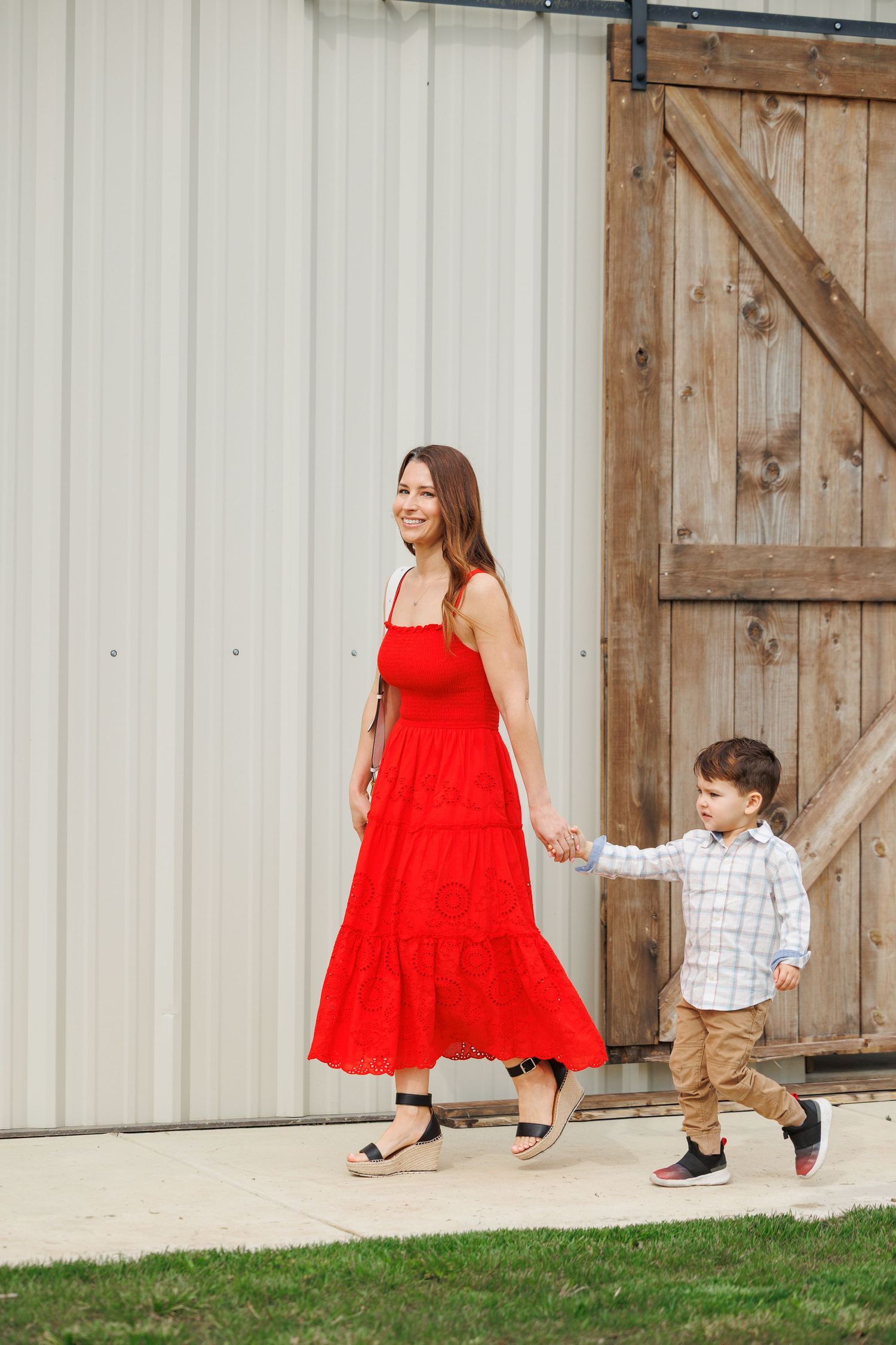 A person in a flowing red dress walks and dances playfully with a small child along a rustic barn wall.
