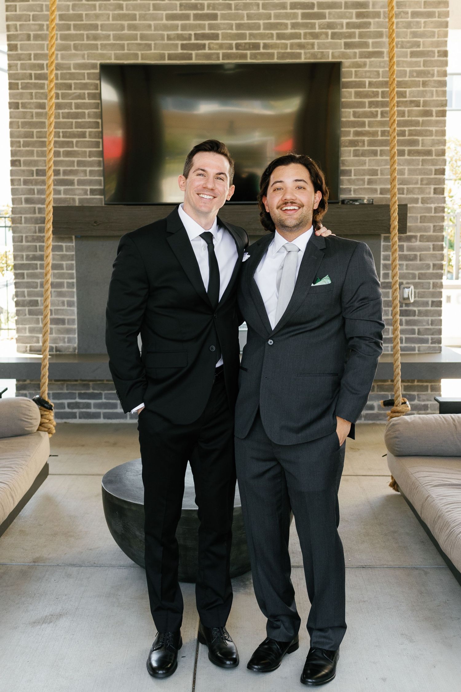 Two people in black suits and ties stand together smiling on a covered patio with brick walls.