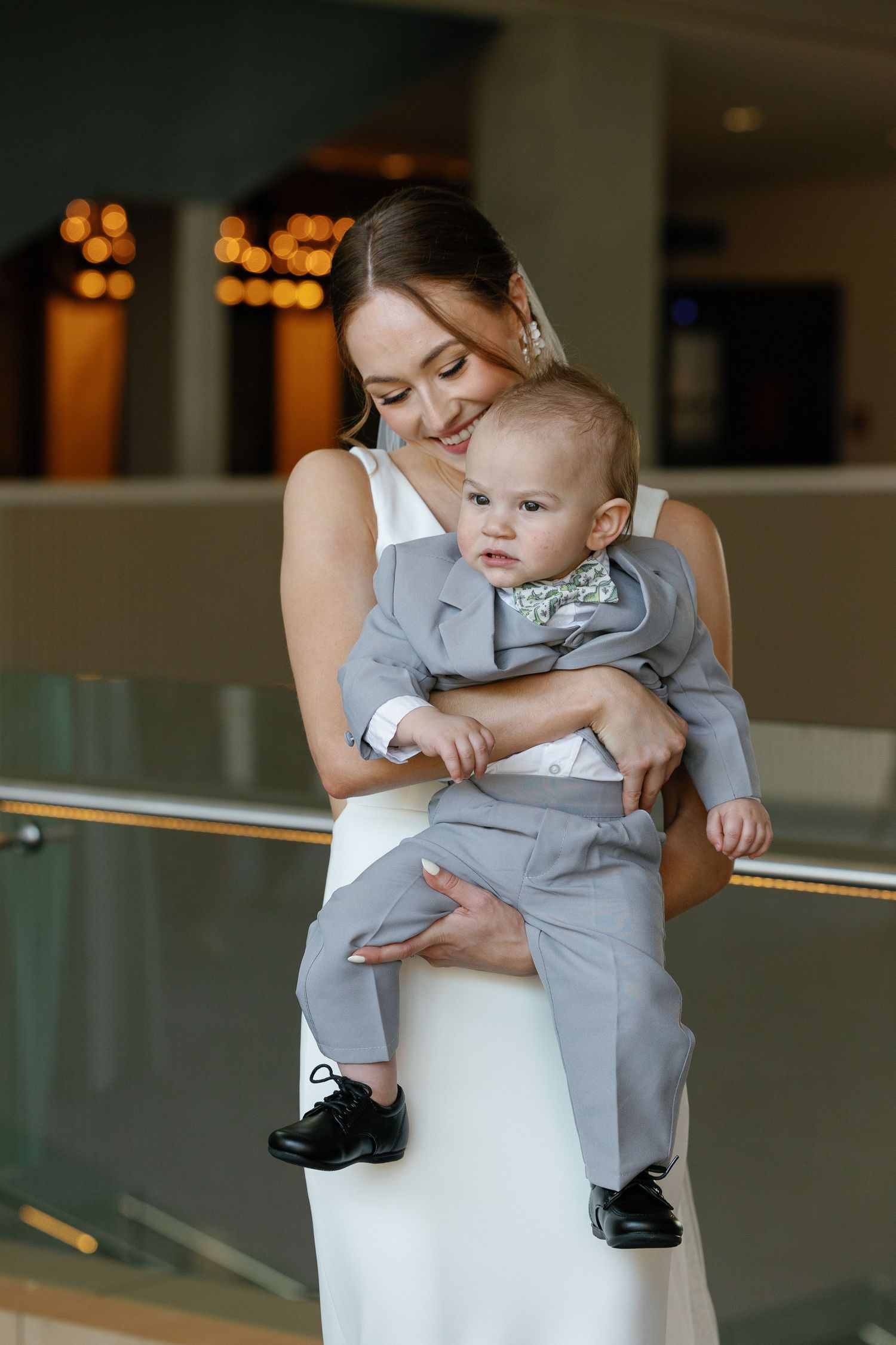 A mother in a white dress holds her baby wearing a gray outfit and dress shoes in an elegant indoor setting.