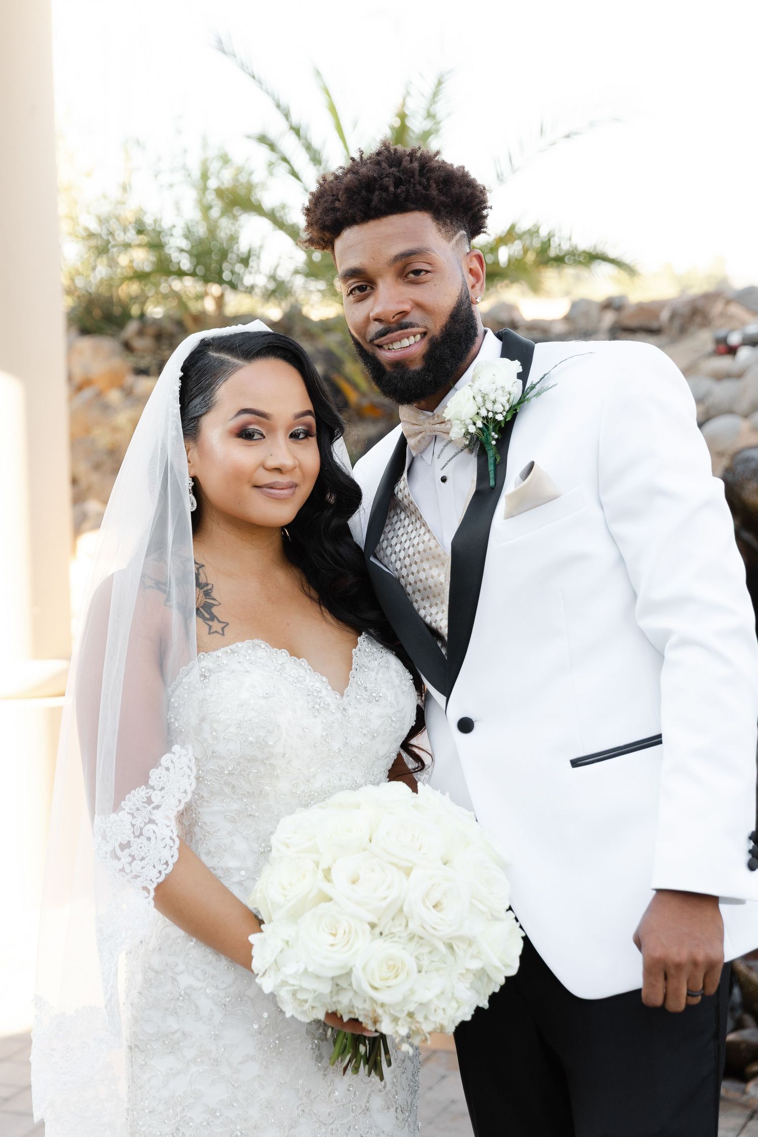 A wedding couple in white formal attire pose together while holding a bouquet of white flowers.