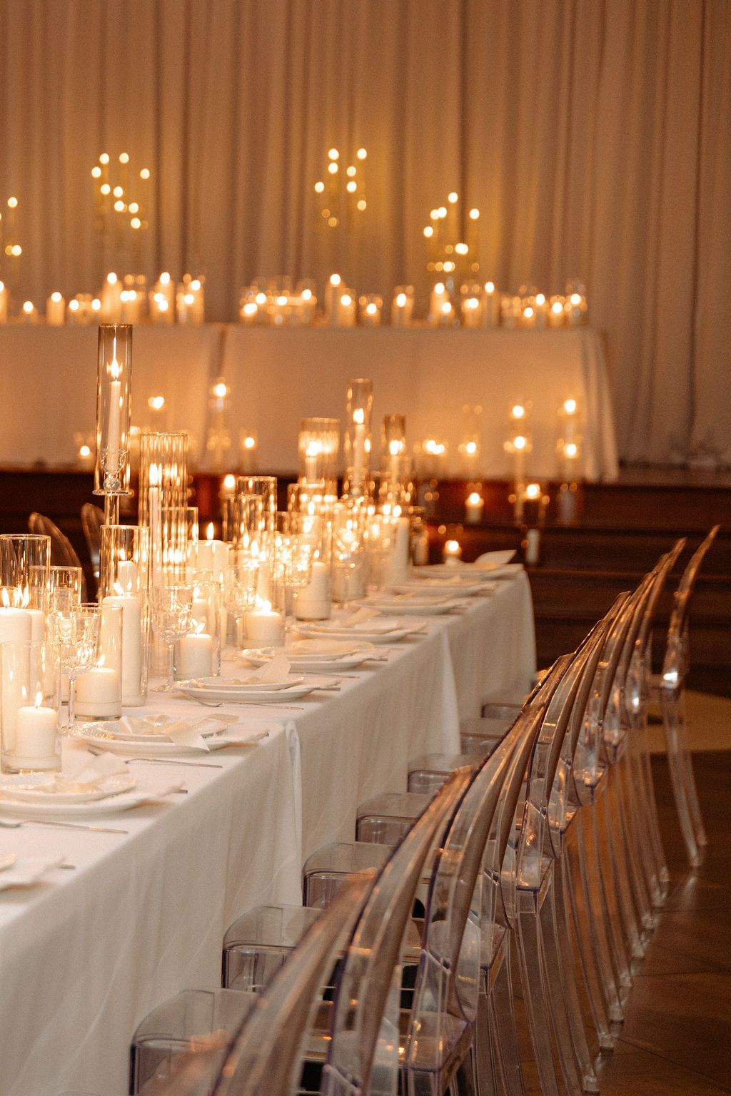 Long banquet table set with elegant white linens, ghost chairs and glowing candles creating a romantic wedding reception atmosphere.