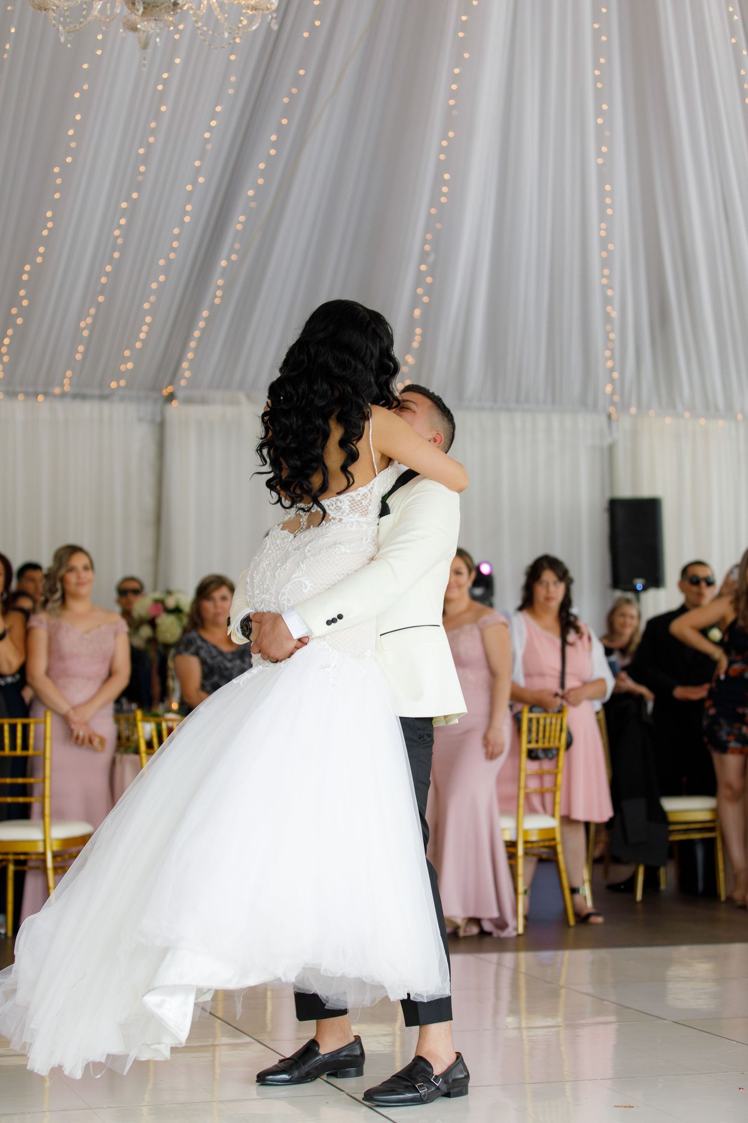 A couple shares their first dance at a wedding reception with bridesmaids in pink dresses watching in the background.