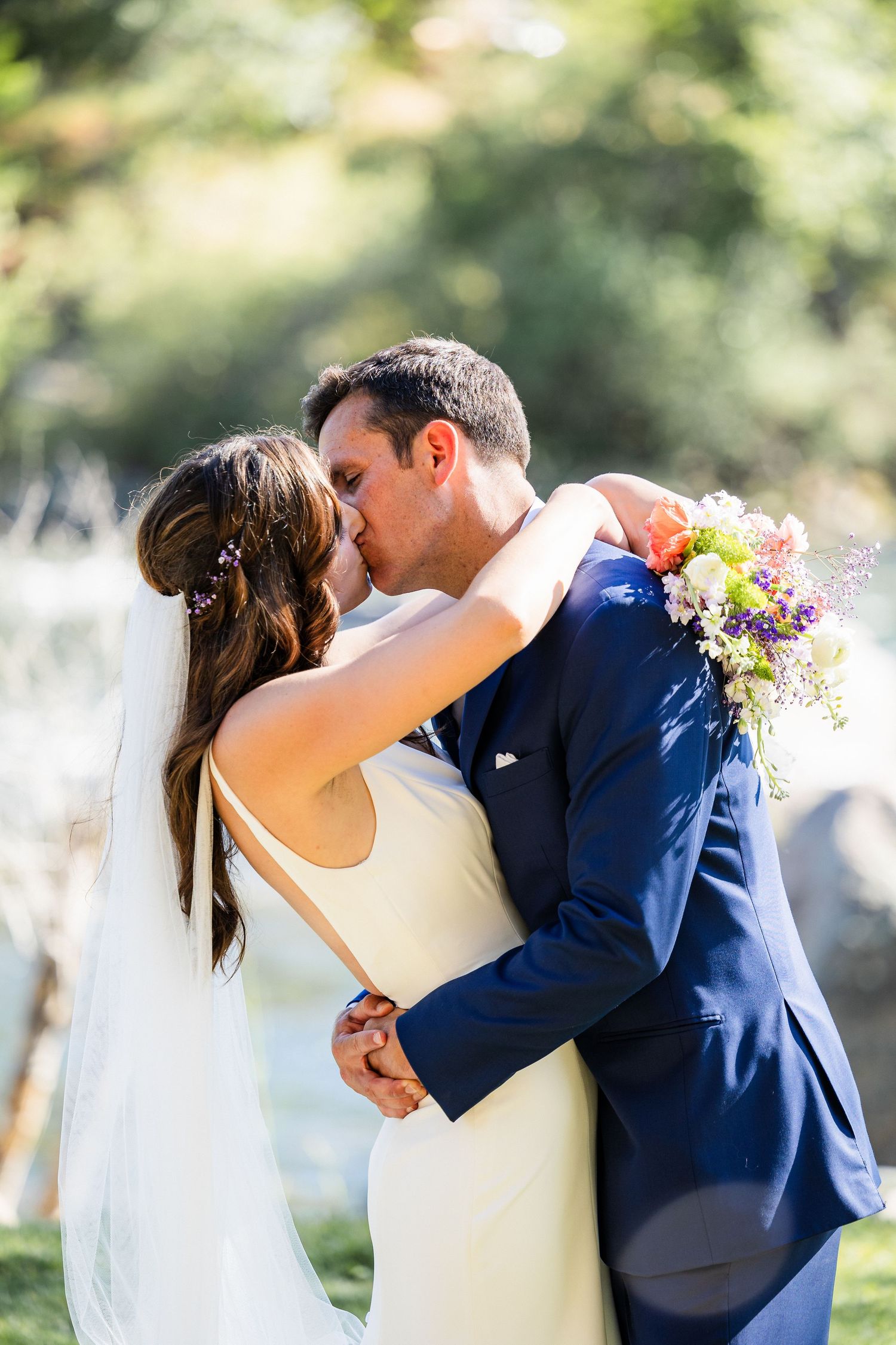 A romantic wedding couple embraces and kisses in a garden setting with soft natural lighting and floral bouquet details.