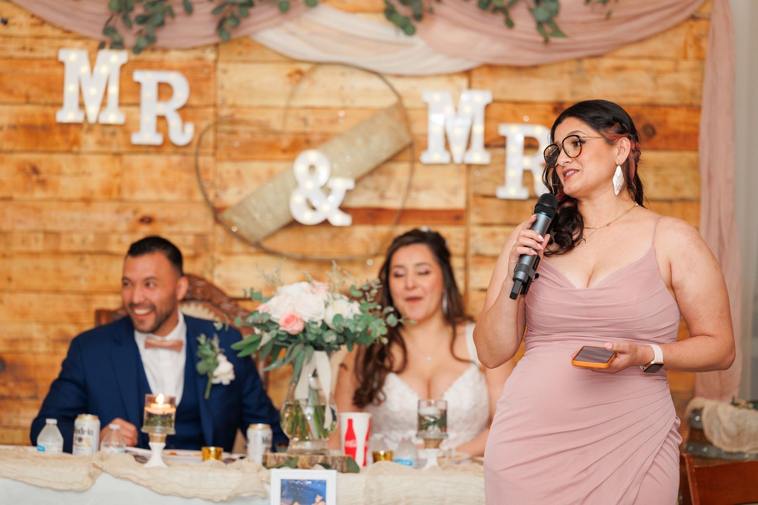 Wedding reception guests seated at long wooden tables inside rustic barn venue with string lights and pink decor.