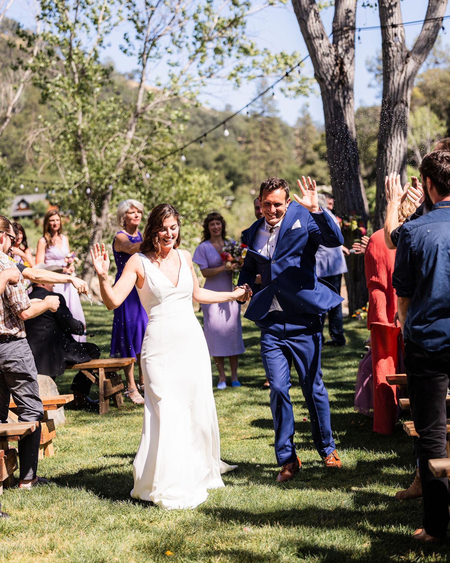 Wedding guests dance and celebrate outdoors in a sunny park setting with trees and greenery in the background.