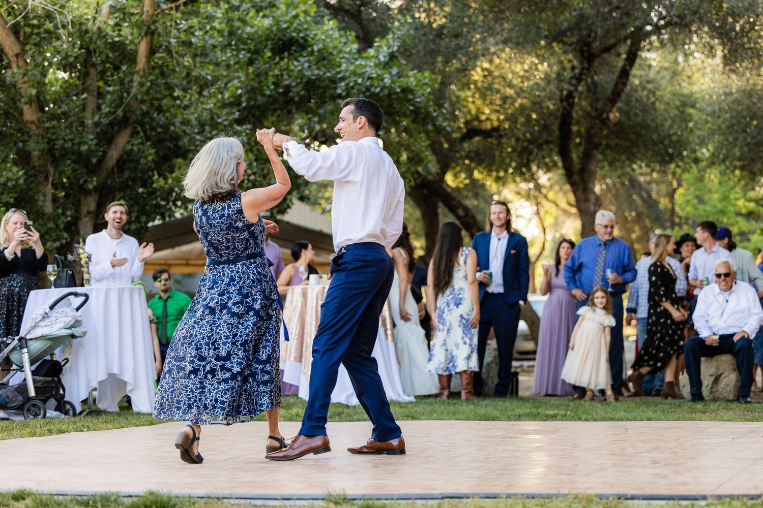 People dancing and celebrating at an outdoor wedding reception under trees during golden hour.