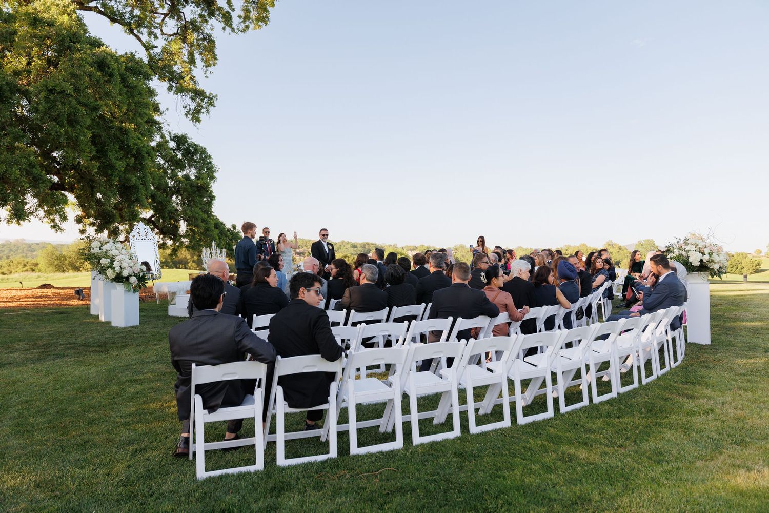 White wedding chairs arranged in rows on a grassy lawn during an outdoor ceremony at sunset.