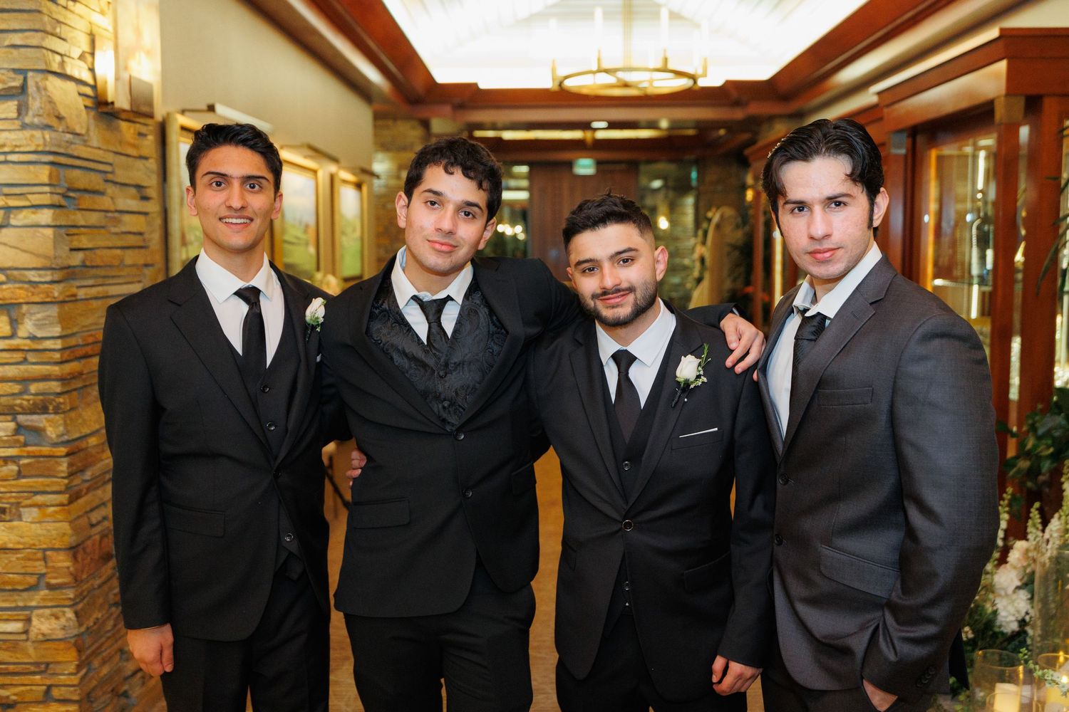 A group wearing black suits and ties stands together in a formal indoor setting with warm lighting and stone walls.