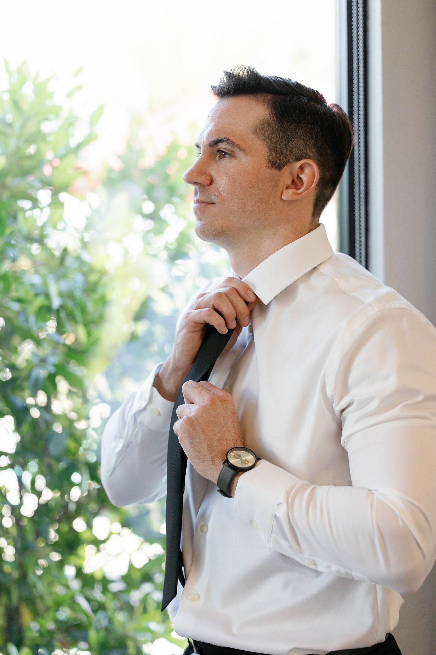 Man in white dress shirt adjusts his tie while standing by a window with natural light and greenery outside.