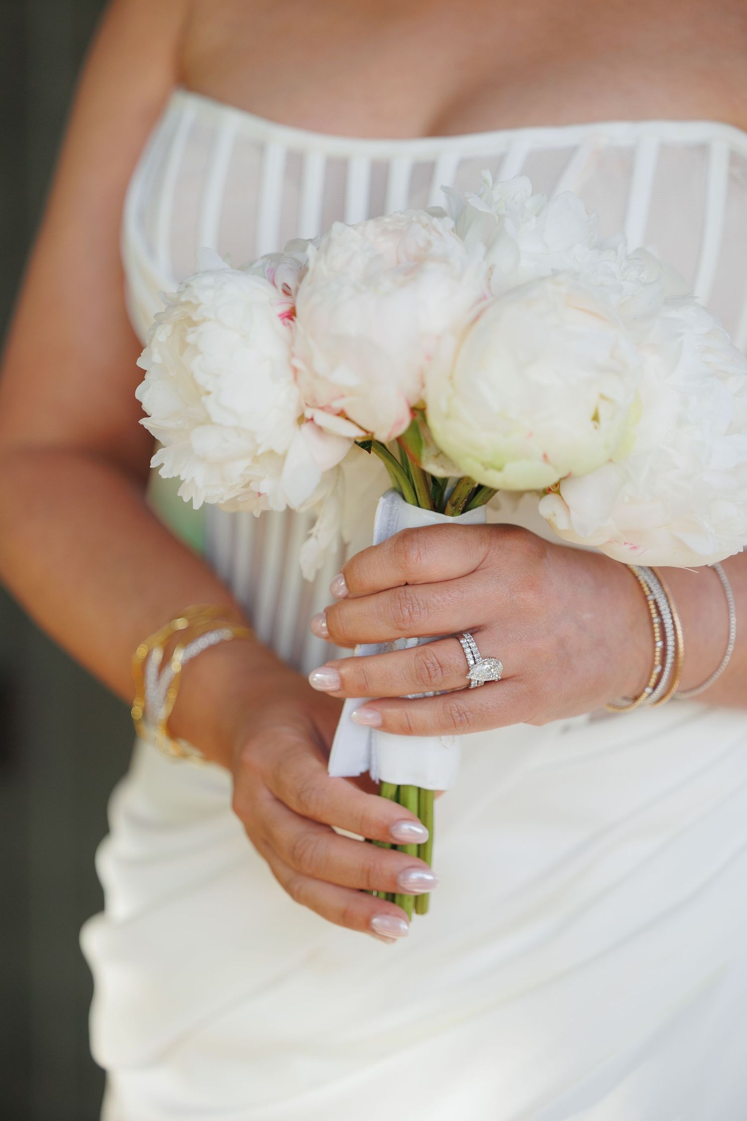 Close-up view of hands holding a white peony bouquet with diamond engagement ring and delicate silver bracelet.