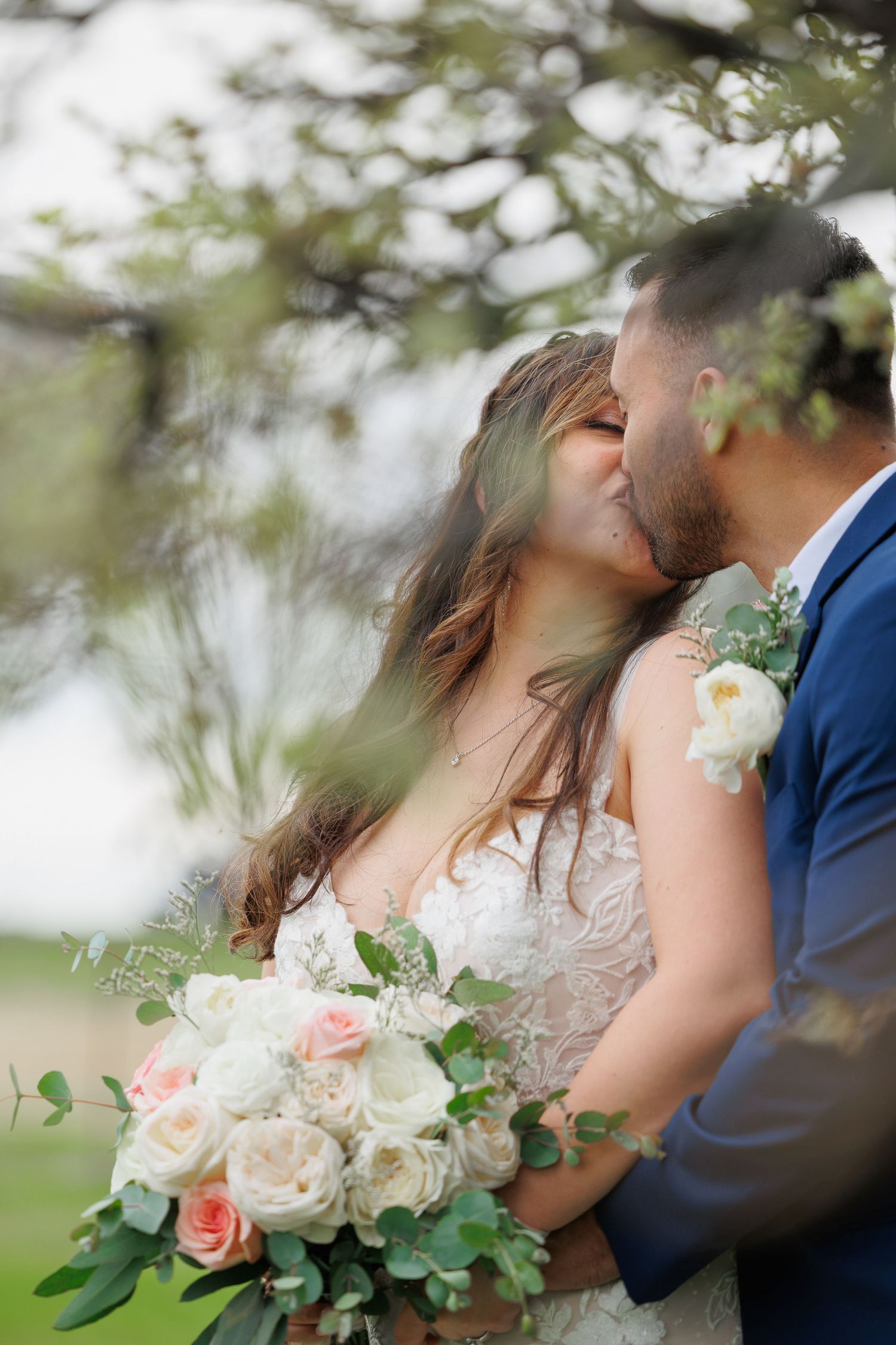 Romantic wedding couple shares intimate moment through blossoming spring branches with soft pink and white floral bouquet.