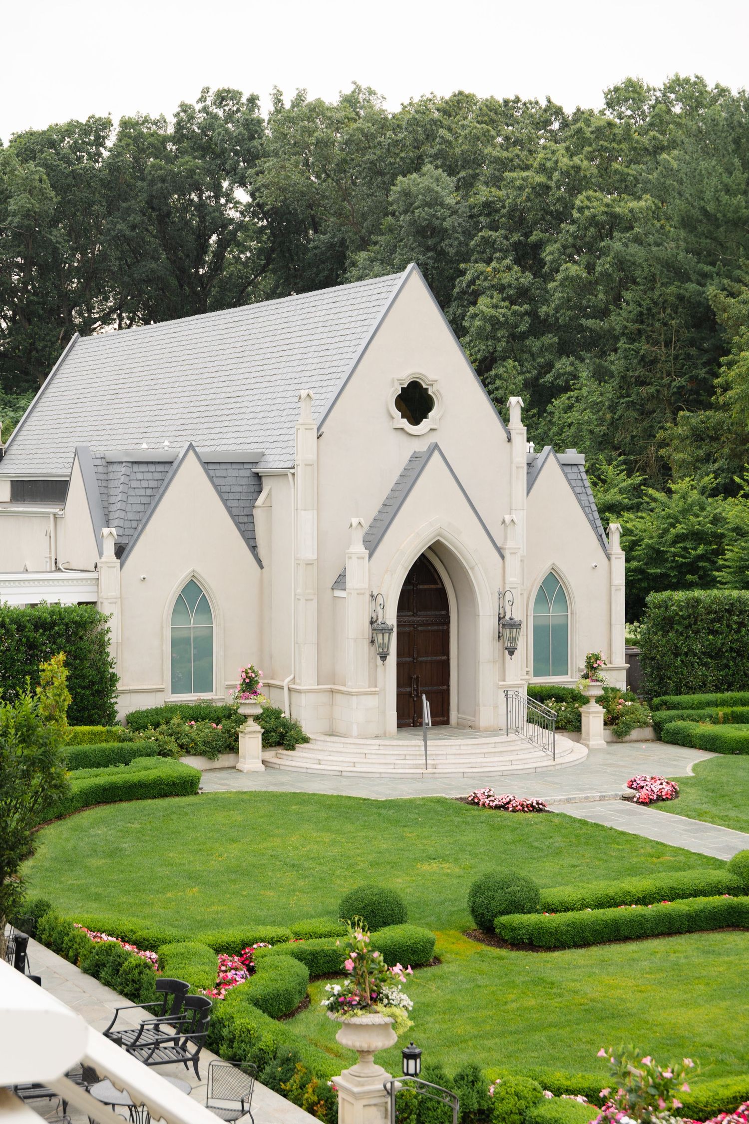 White gothic-style chapel with pointed arches and green shutters sits nestled among manicured hedges and lawns.