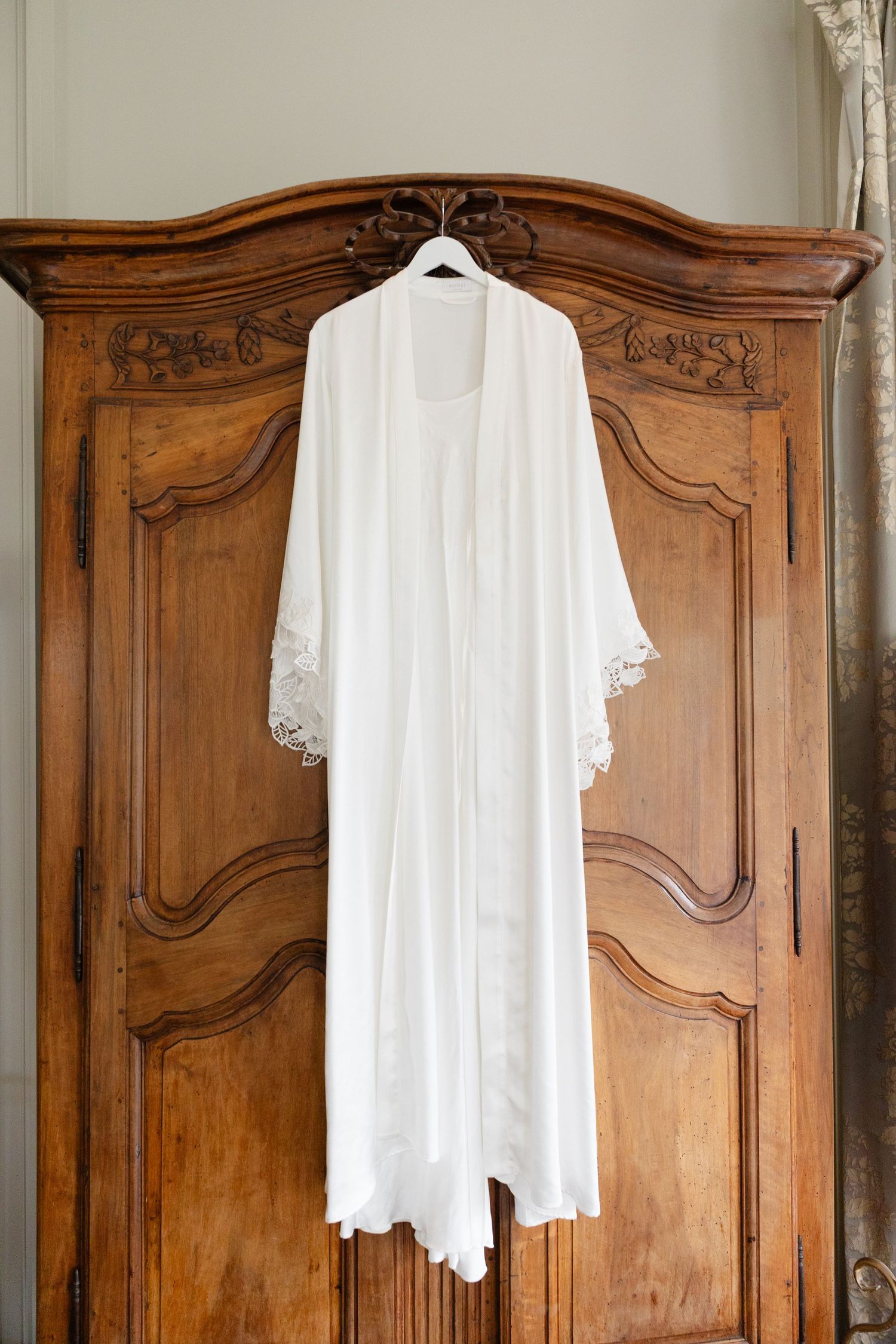 Elegant white lace bridal robe hanging on antique wooden armoire with ornate carved details.