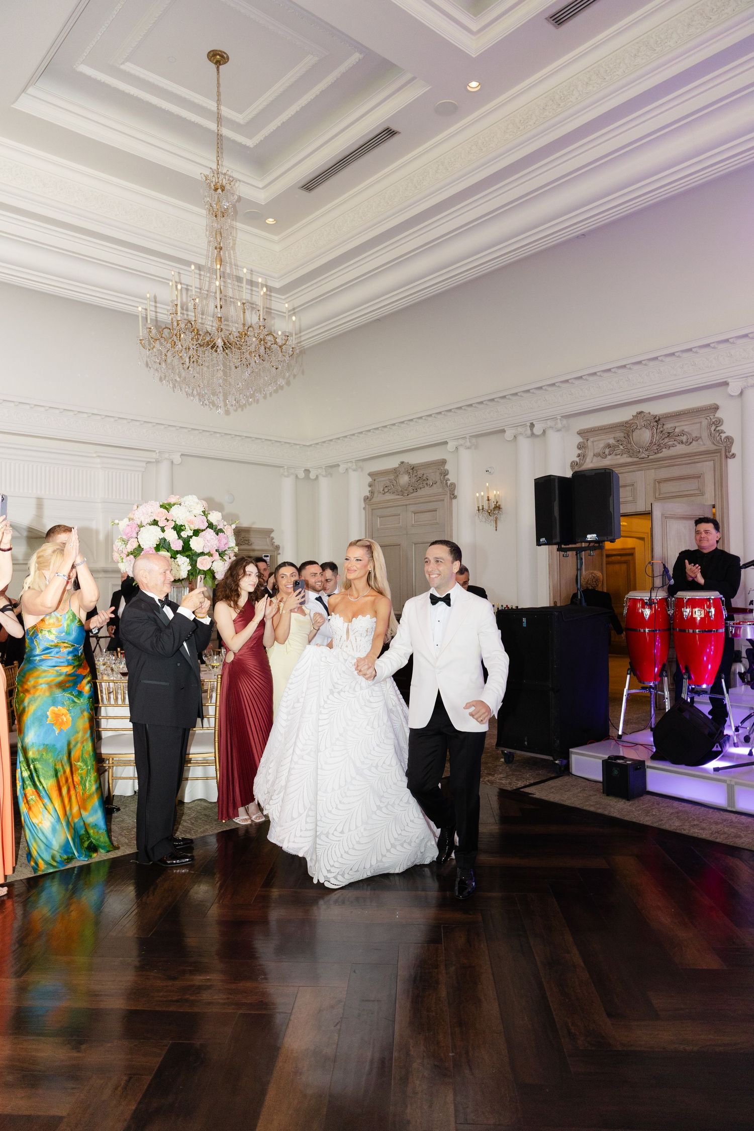 Wedding guests dance together in an elegant ballroom with crystal chandeliers and white ornate moldings.