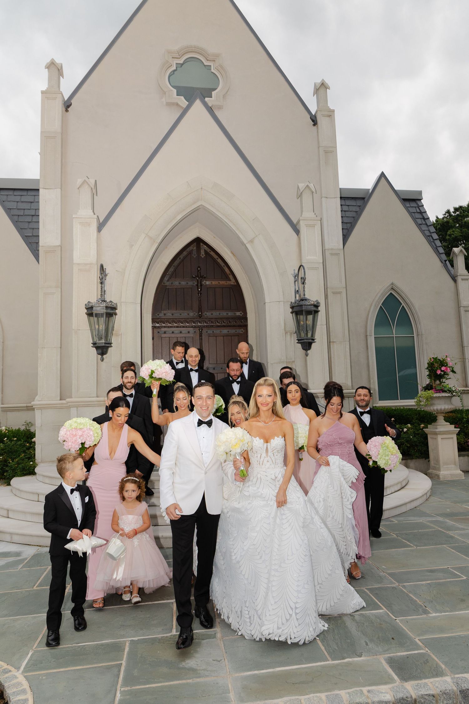 Wedding party poses playfully in front of a white Gothic church with pointed arches and lanterns.