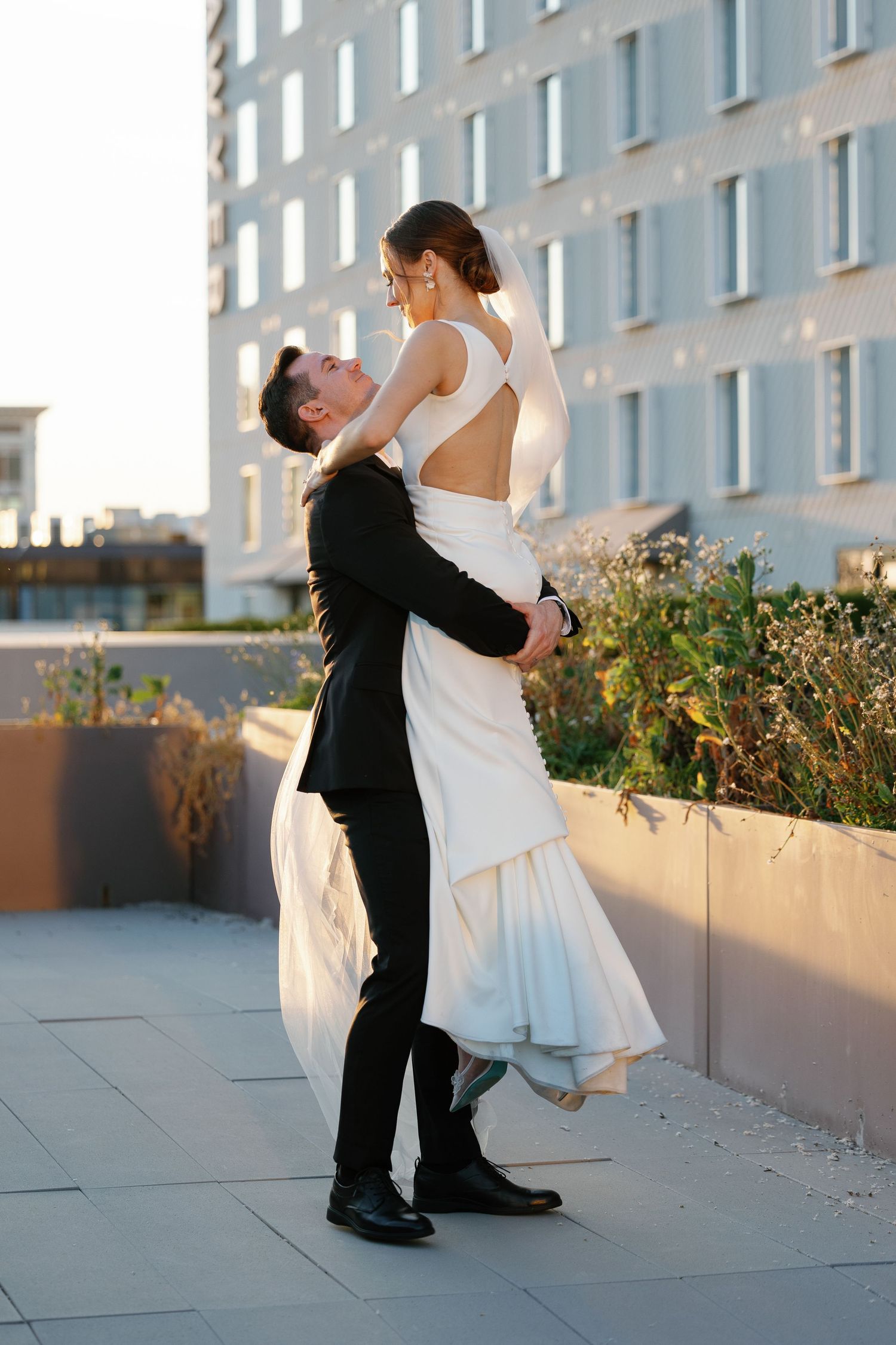 A romantic wedding couple embraces on a rooftop terrace with a modern building backdrop at sunset.
