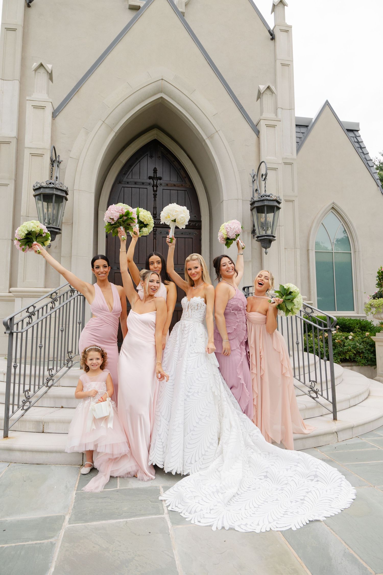 Bride in white gown with bridesmaids in pink dresses celebrating outside a church, holding bouquets aloft.