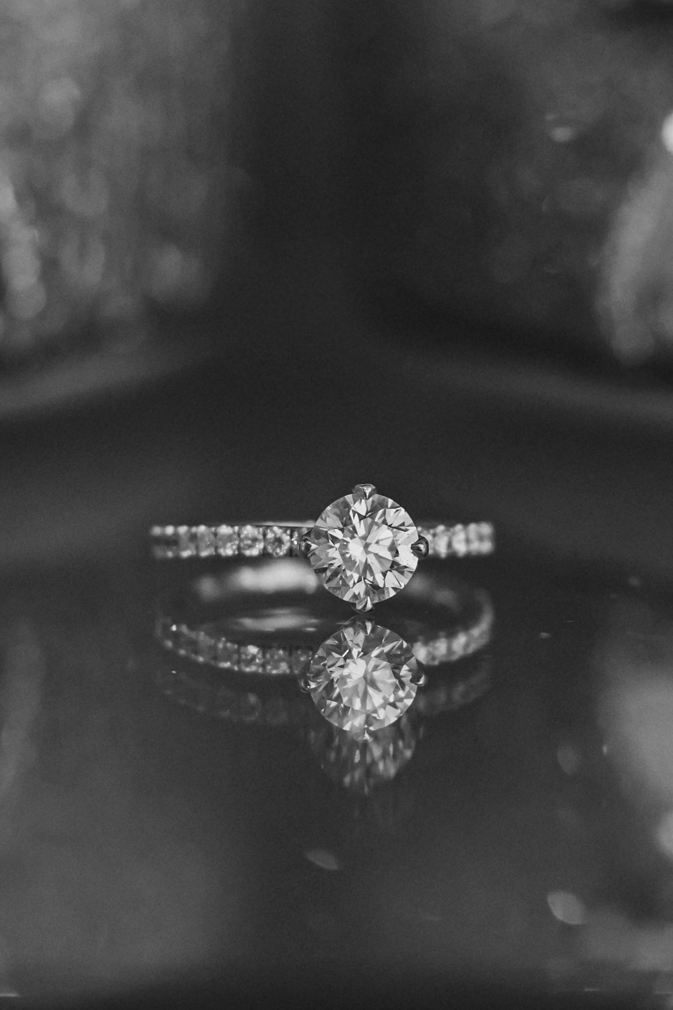 Close-up black and white photo of an elegant diamond engagement ring with a solitaire center stone and pave band.