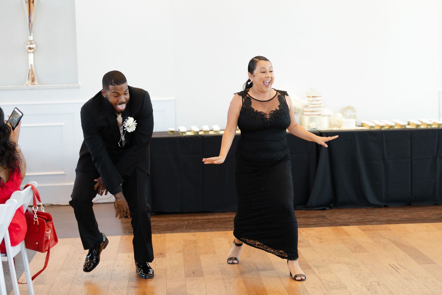 Two people in formal black attire dance energetically on a wooden floor at an event with a buffet table in the background.
