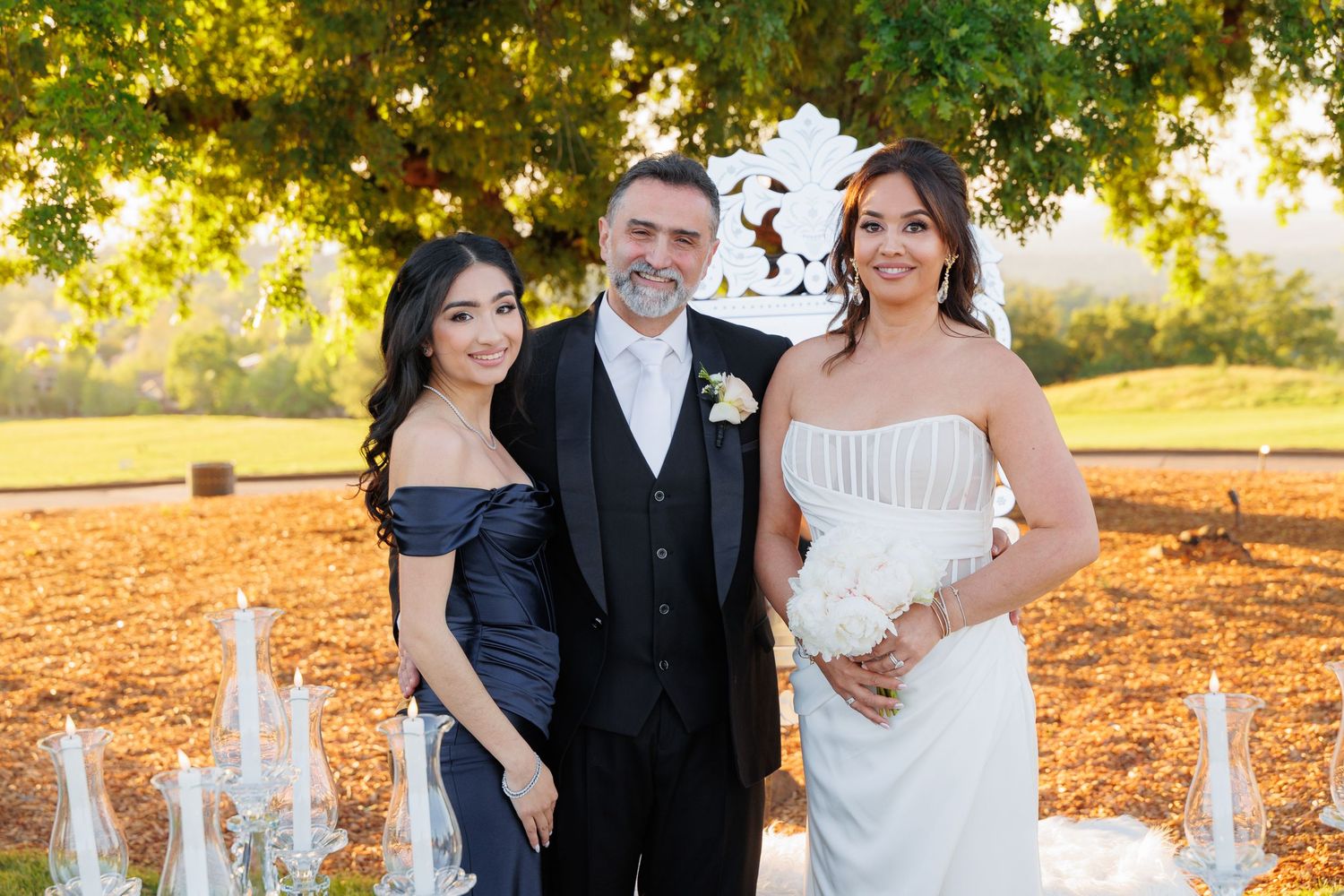 A wedding party poses outdoors during golden hour under trees with warm sunlight filtering through the branches.