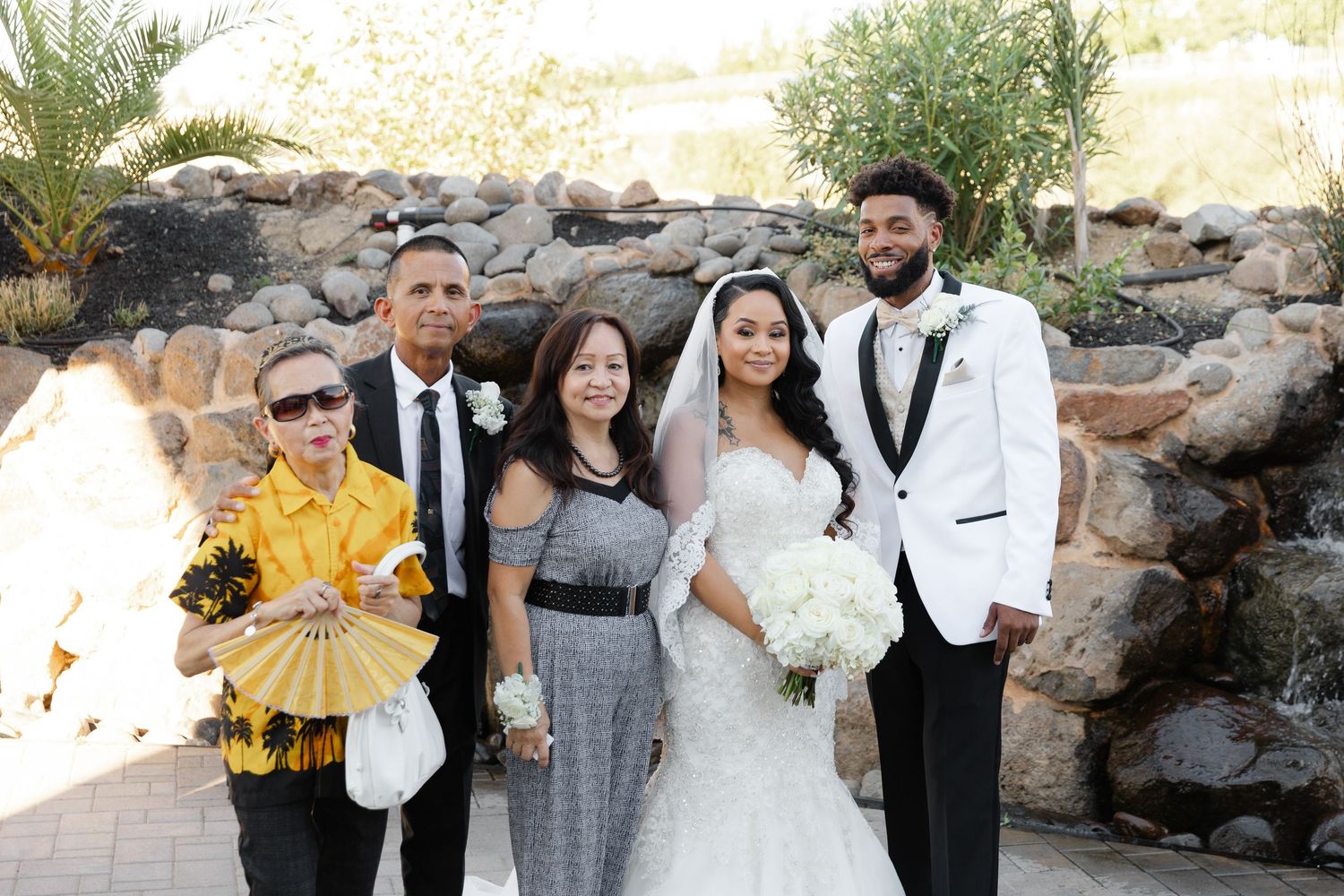 Wedding party members pose together outdoors in formal attire near a stone wall on a sunny day.